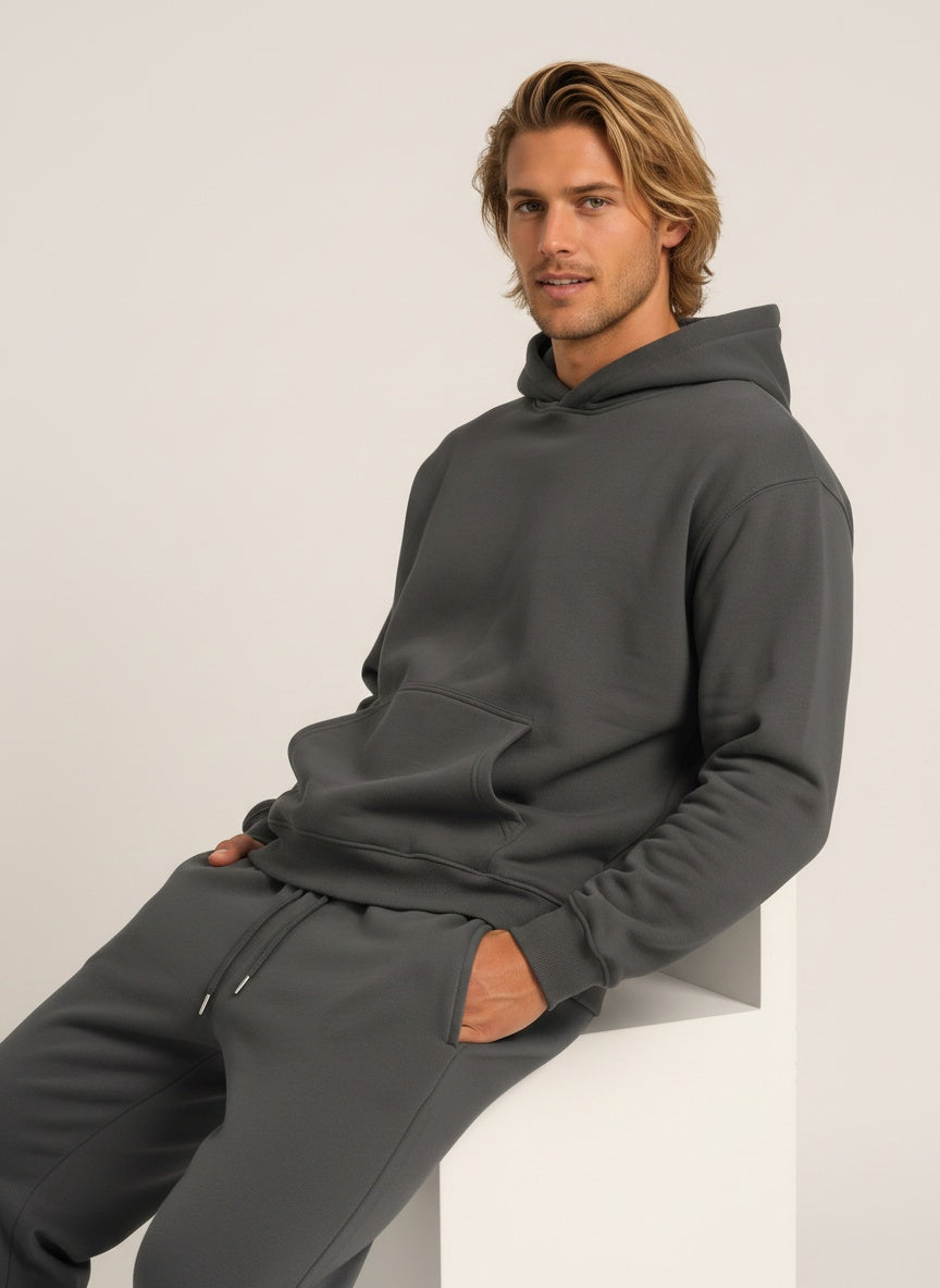 A man wearing a dark gray matching lounge set consisting of a pullover hoodie and sweatpants, sitting on a white geometric block against a light gray studio background.