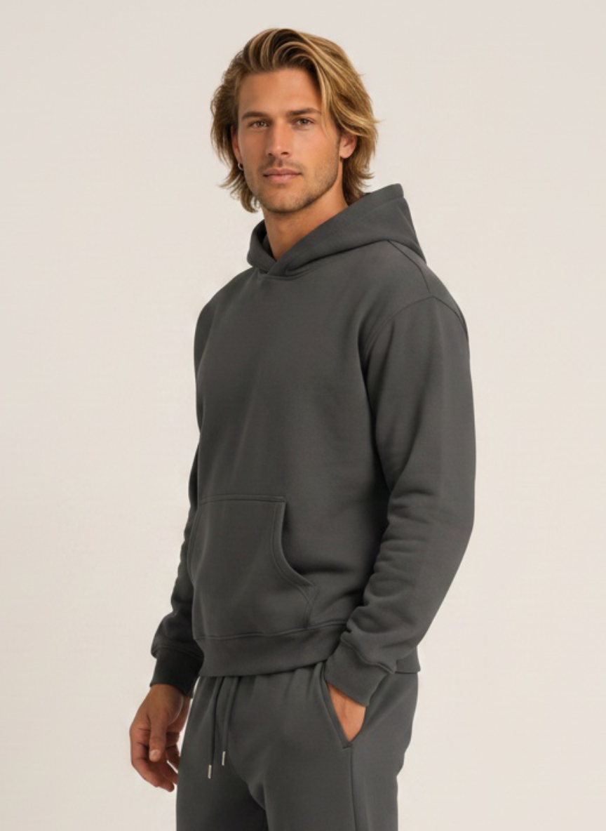 A man wearing a dark gray pullover hoodie and matching sweatpants, standing against a plain light beige studio background.