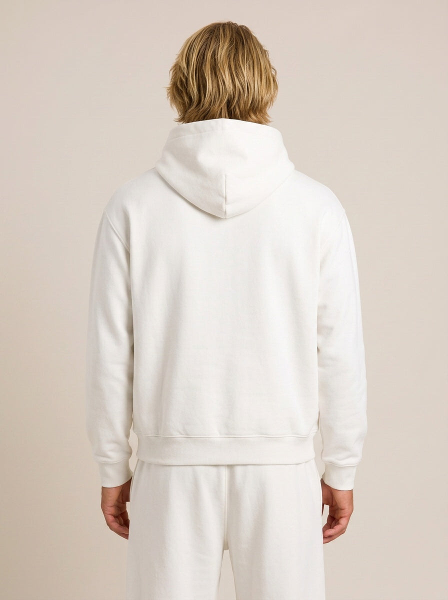 A man wearing a dark gray matching lounge set consisting of a hooded sweatshirt and jogger pants, standing in profile against a solid beige studio background.