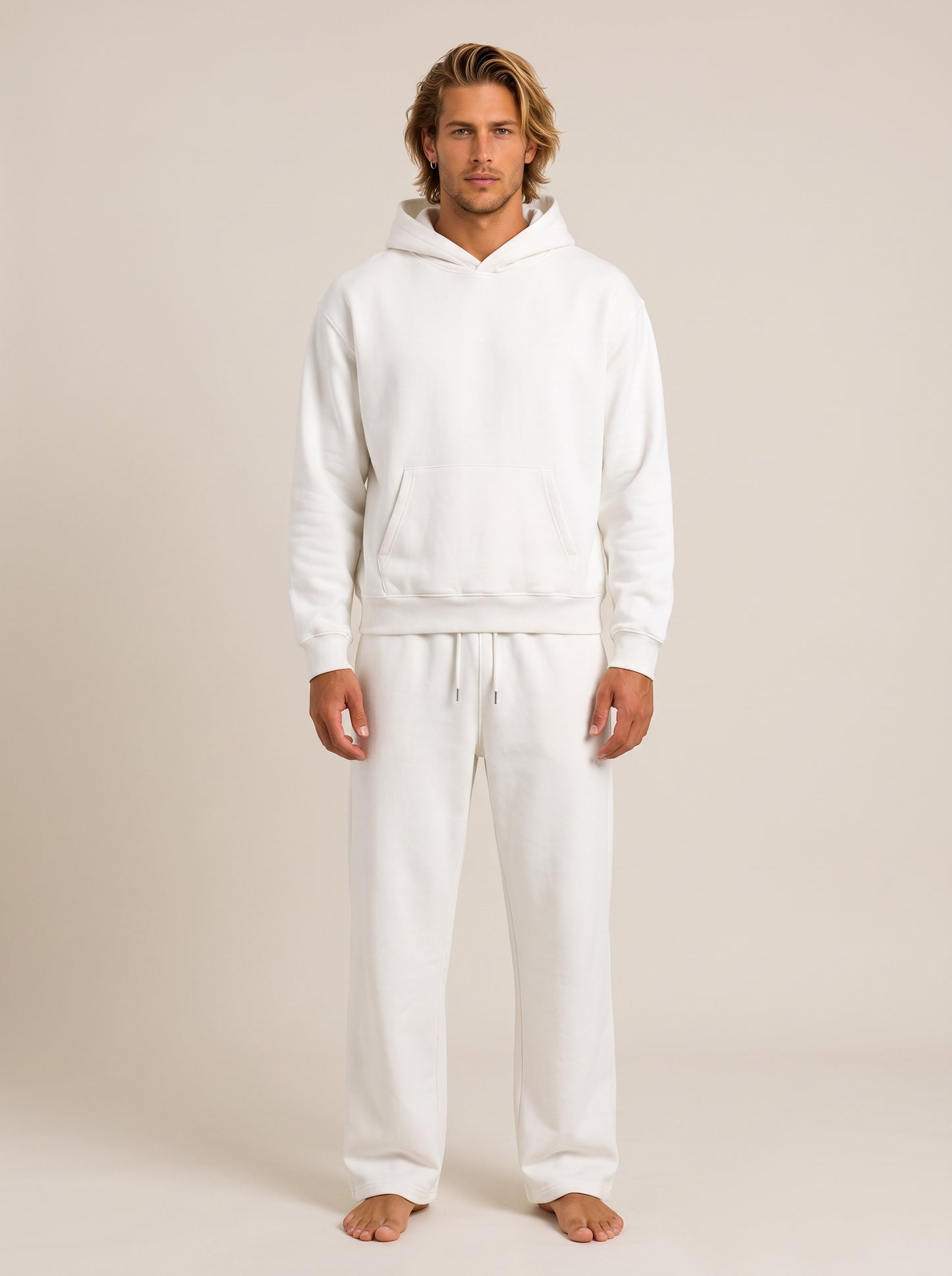 A man modeling a matching white loungewear set consisting of a pullover hoodie and sweatpants, standing against a solid light beige studio background.