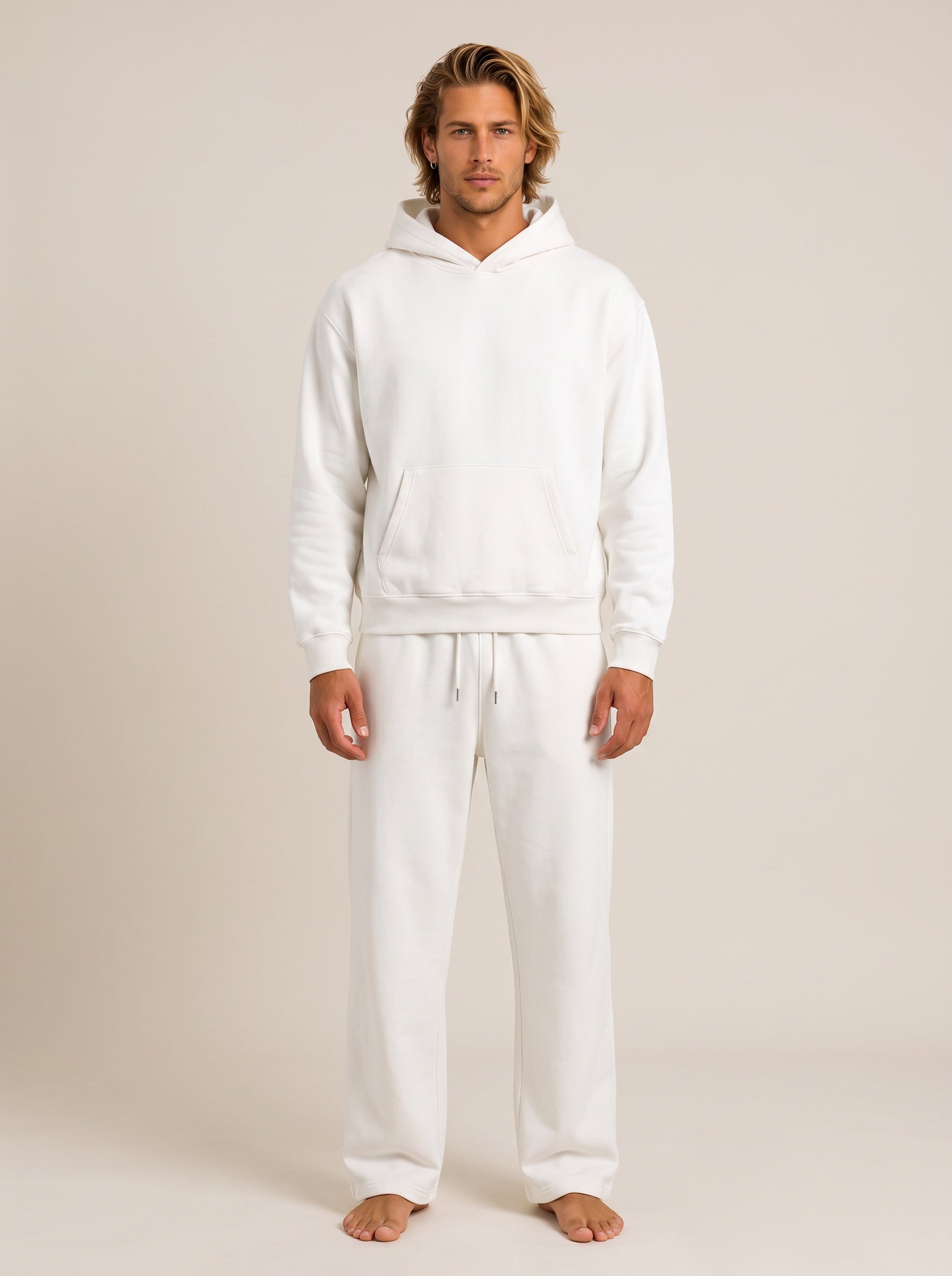 A man modeling a matching white loungewear set consisting of a pullover hoodie and sweatpants, standing against a solid light beige studio background.