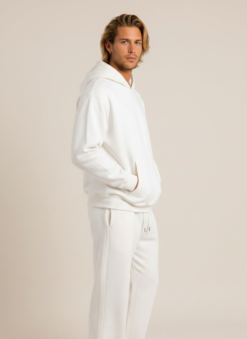 A man wearing a matching white hoodie and sweatpants set, standing in profile against a plain beige studio background.