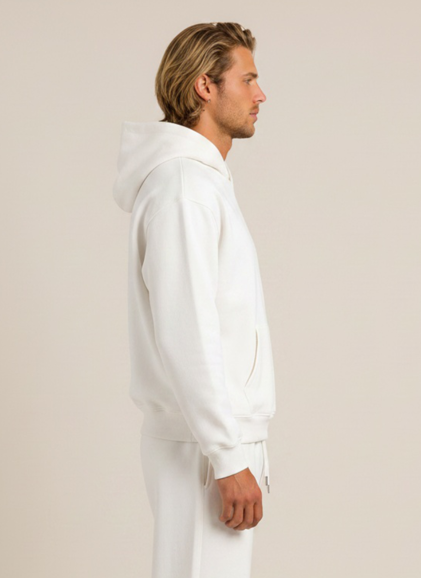 A man in profile wearing a white hooded sweatshirt and matching white sweatpants against a plain beige studio background.