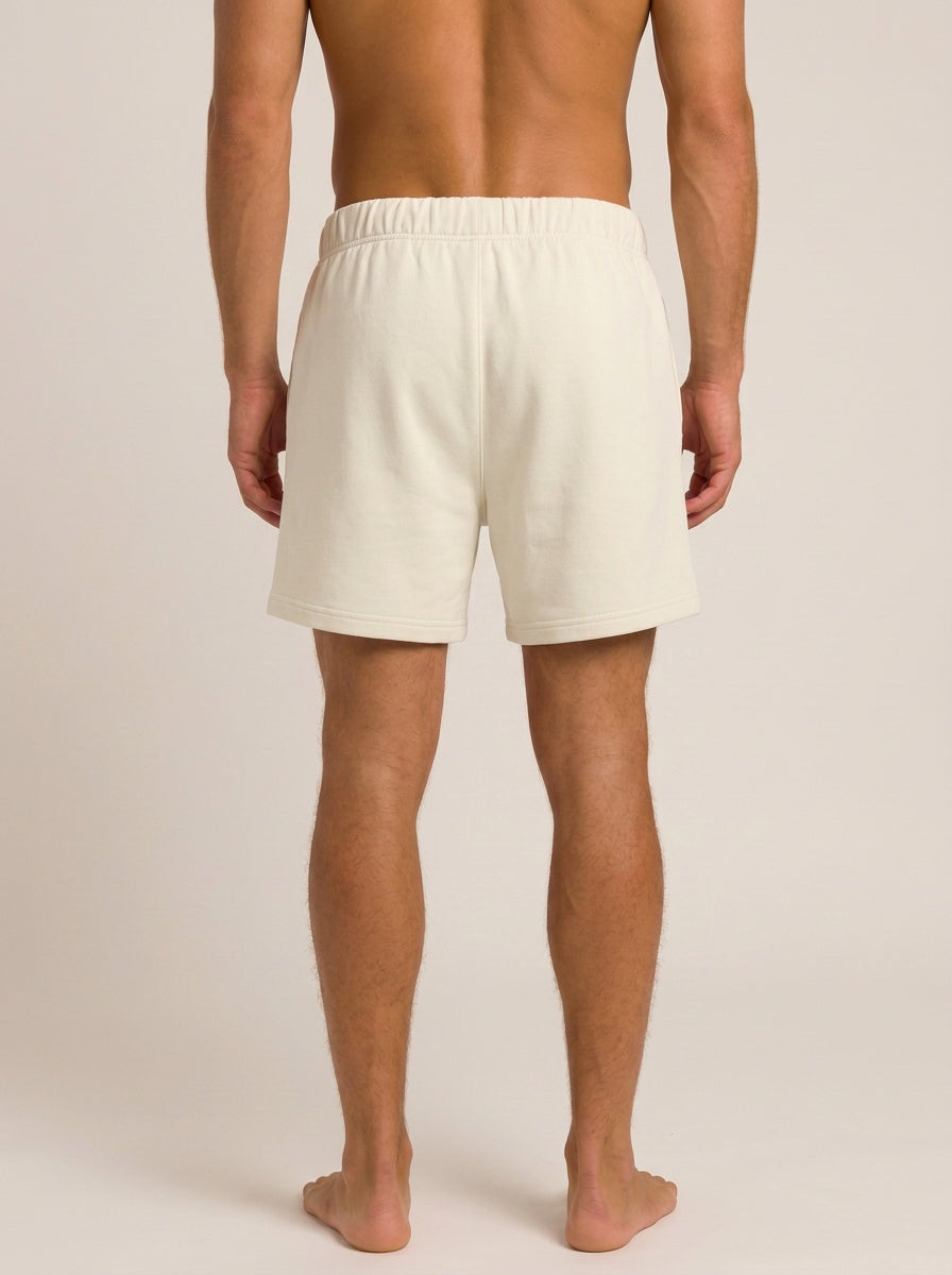 A man shown from the back wearing off-white or cream-colored lounge shorts, standing barefoot against a plain light beige studio background.
