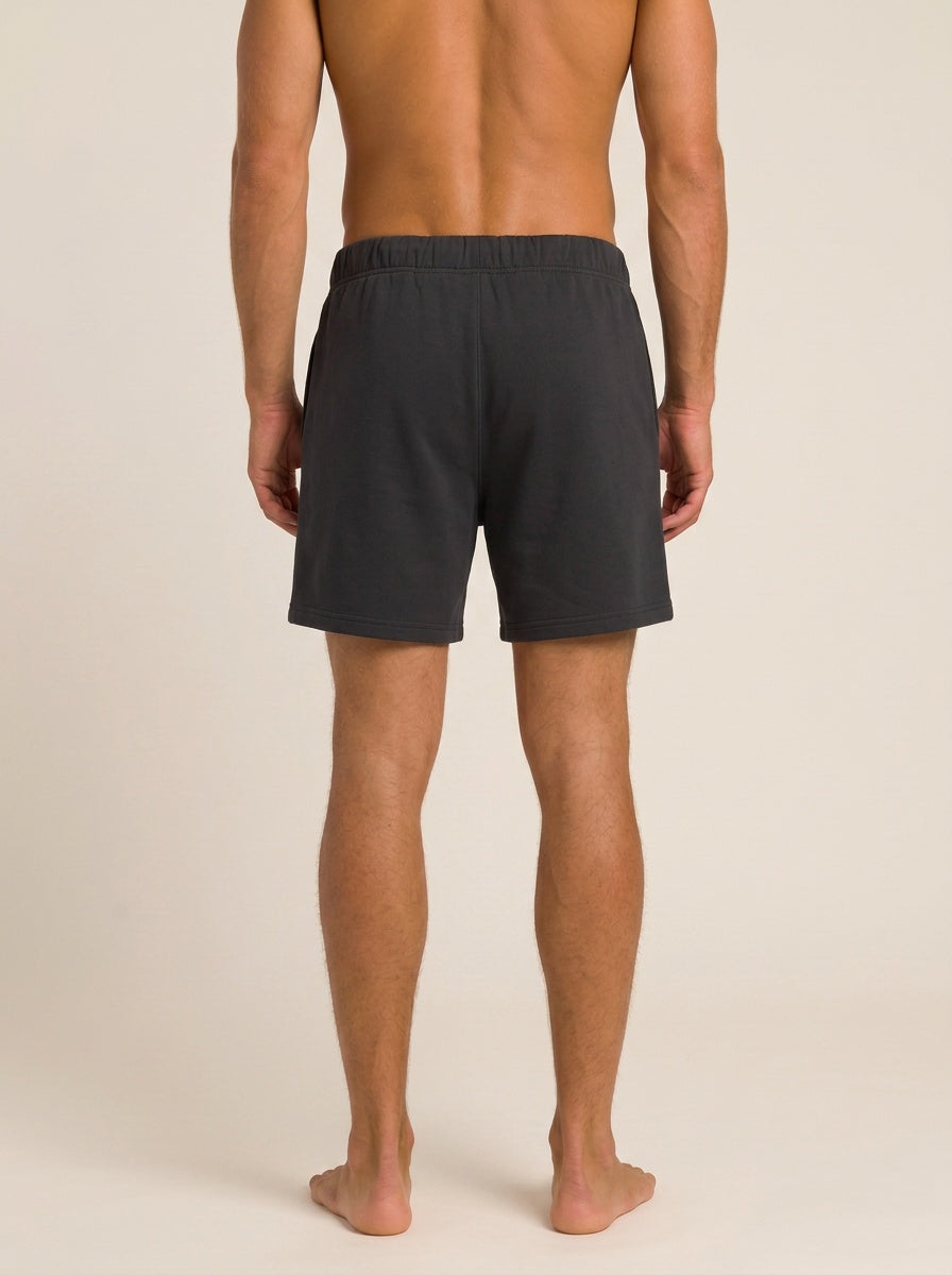Rear view of a man wearing dark charcoal gray athletic shorts against a plain beige studio background.
