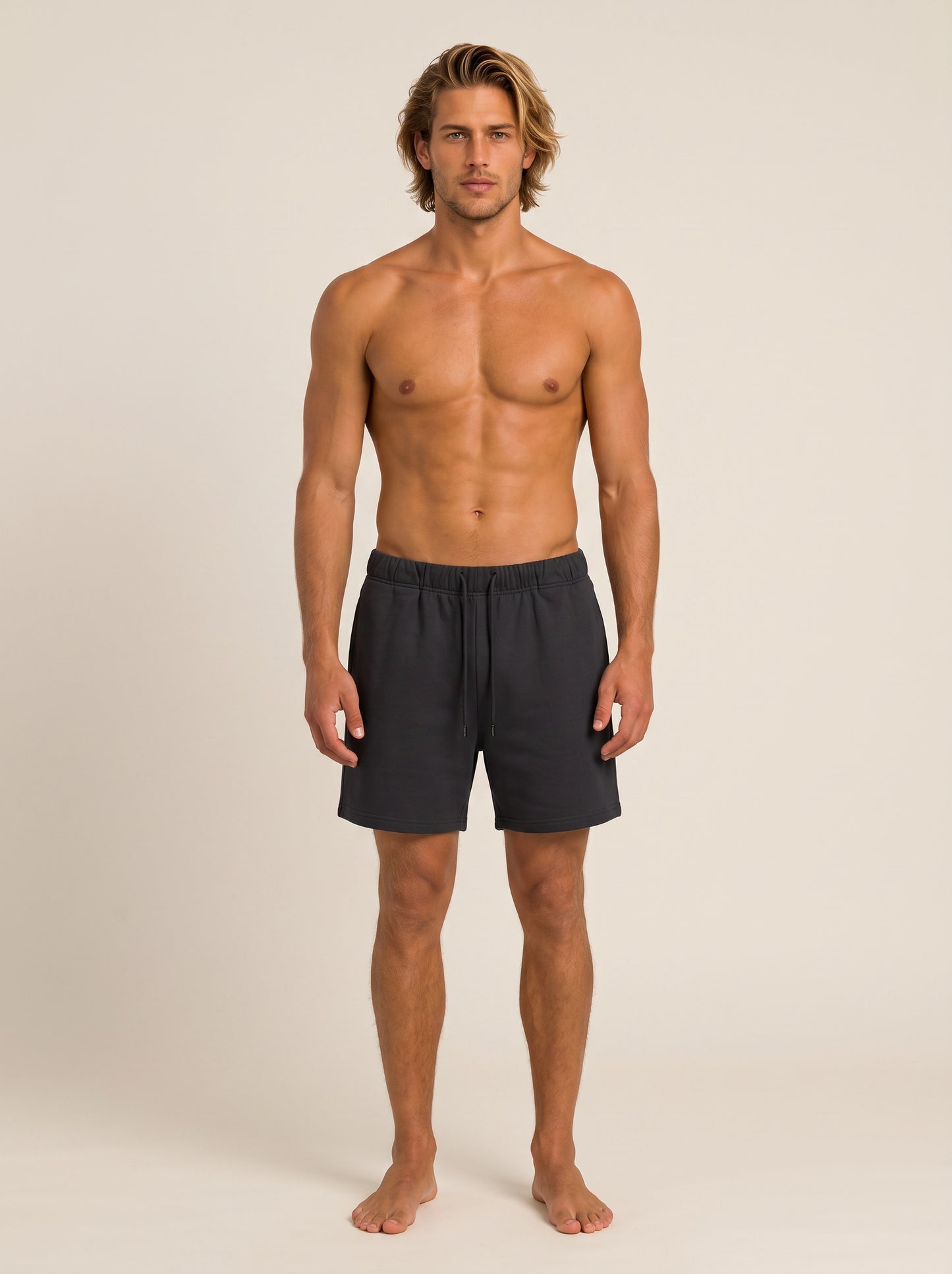 A shirtless male model stands barefoot against a plain warm beige studio background, wearing dark charcoal gray swim shorts.