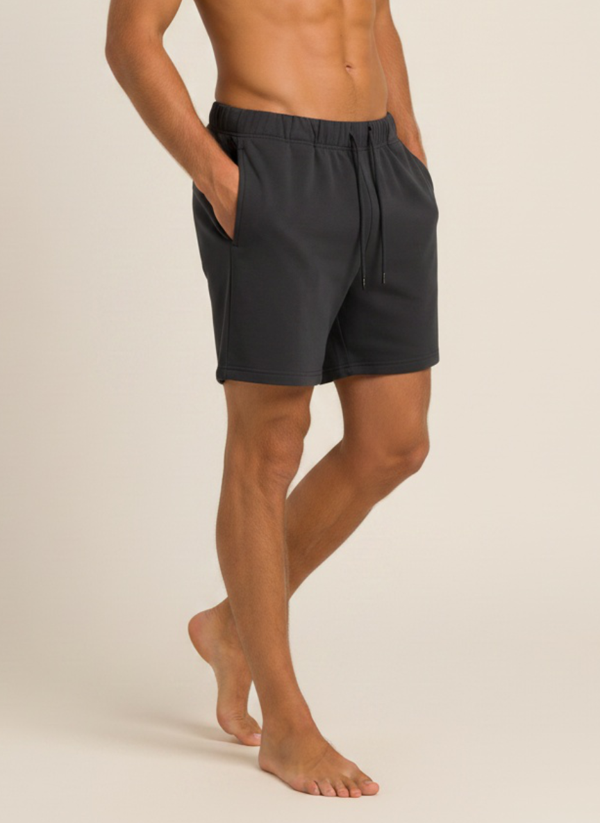 A man wearing dark charcoal gray drawstring shorts, shown from the waist down against a plain light beige studio background.