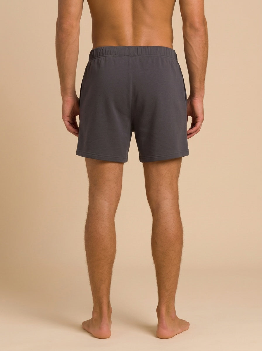 A man shown from the back wearing dark gray athletic shorts, standing barefoot against a solid beige studio background.