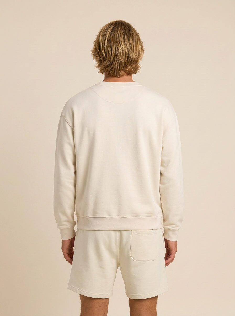 A man viewed from behind wearing a cream-colored crewneck sweatshirt and matching shorts against a solid beige background.