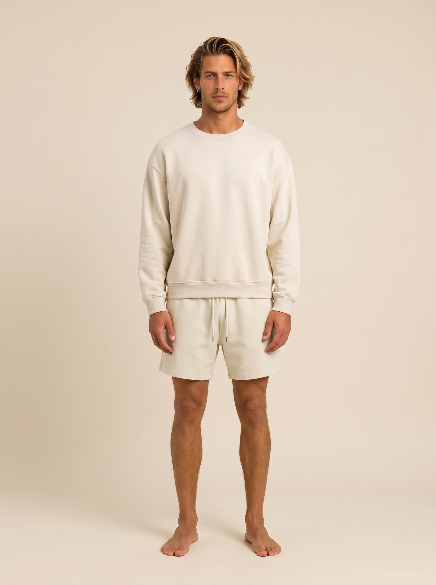 A man stands barefoot against a plain warm beige studio background, wearing a matching cream sweatshirt and shorts set.