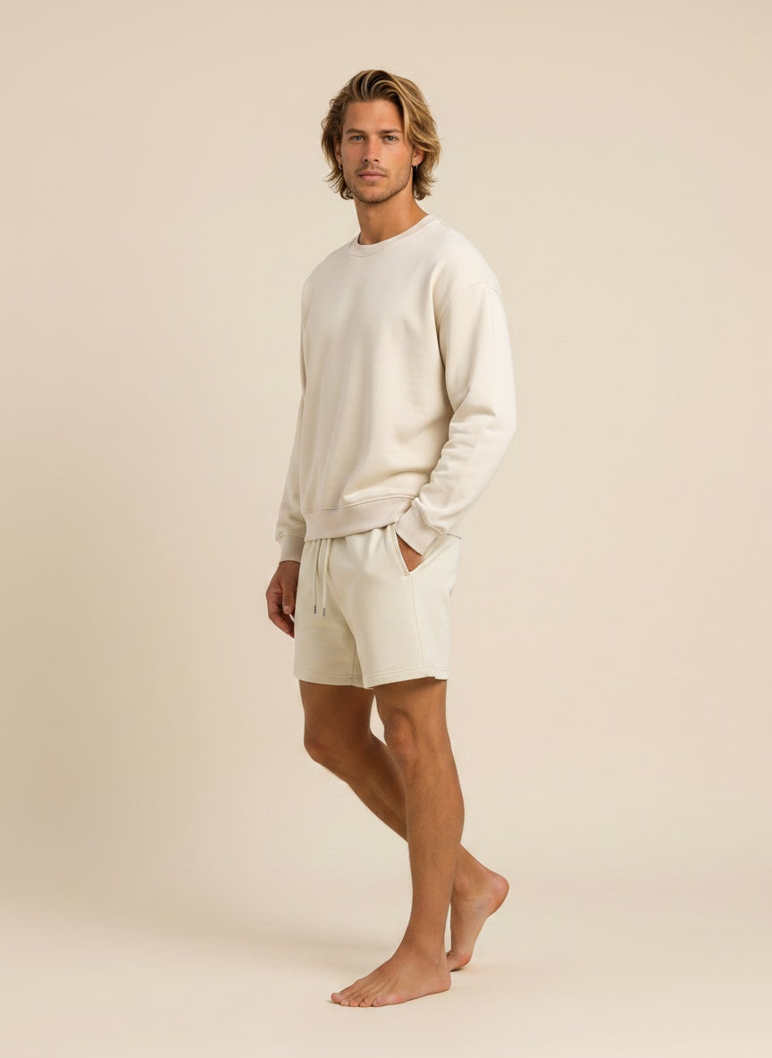 A man modeling a matching cream-colored lounge set consisting of a long-sleeve crewneck sweatshirt and drawstring shorts, standing against a solid light beige studio background.