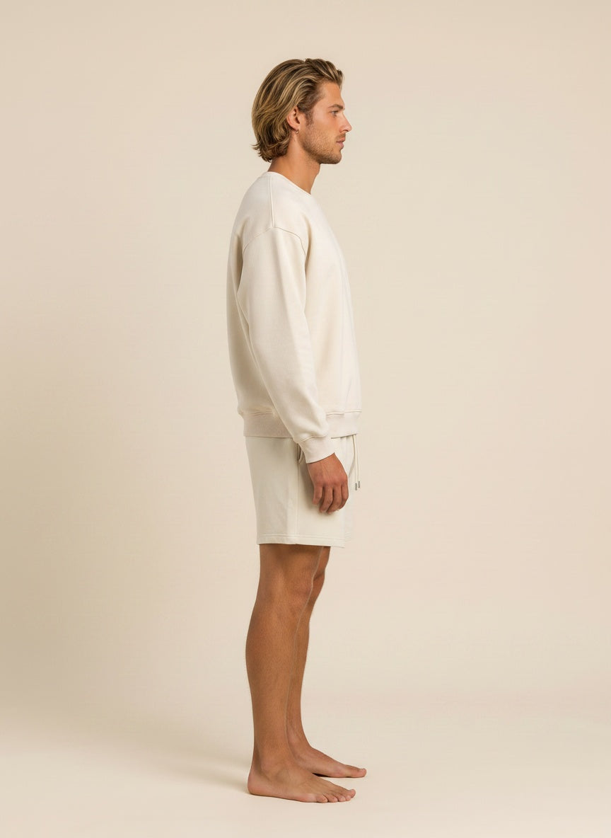 A man in side profile wearing a cream-colored crewneck sweatshirt and matching shorts against a solid beige studio background.