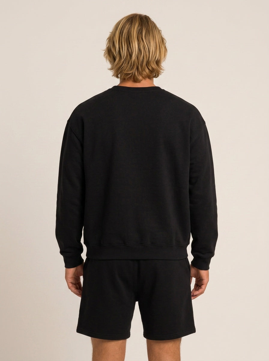 A man viewed from behind wearing a black crewneck sweatshirt and matching black shorts against a plain beige studio background.