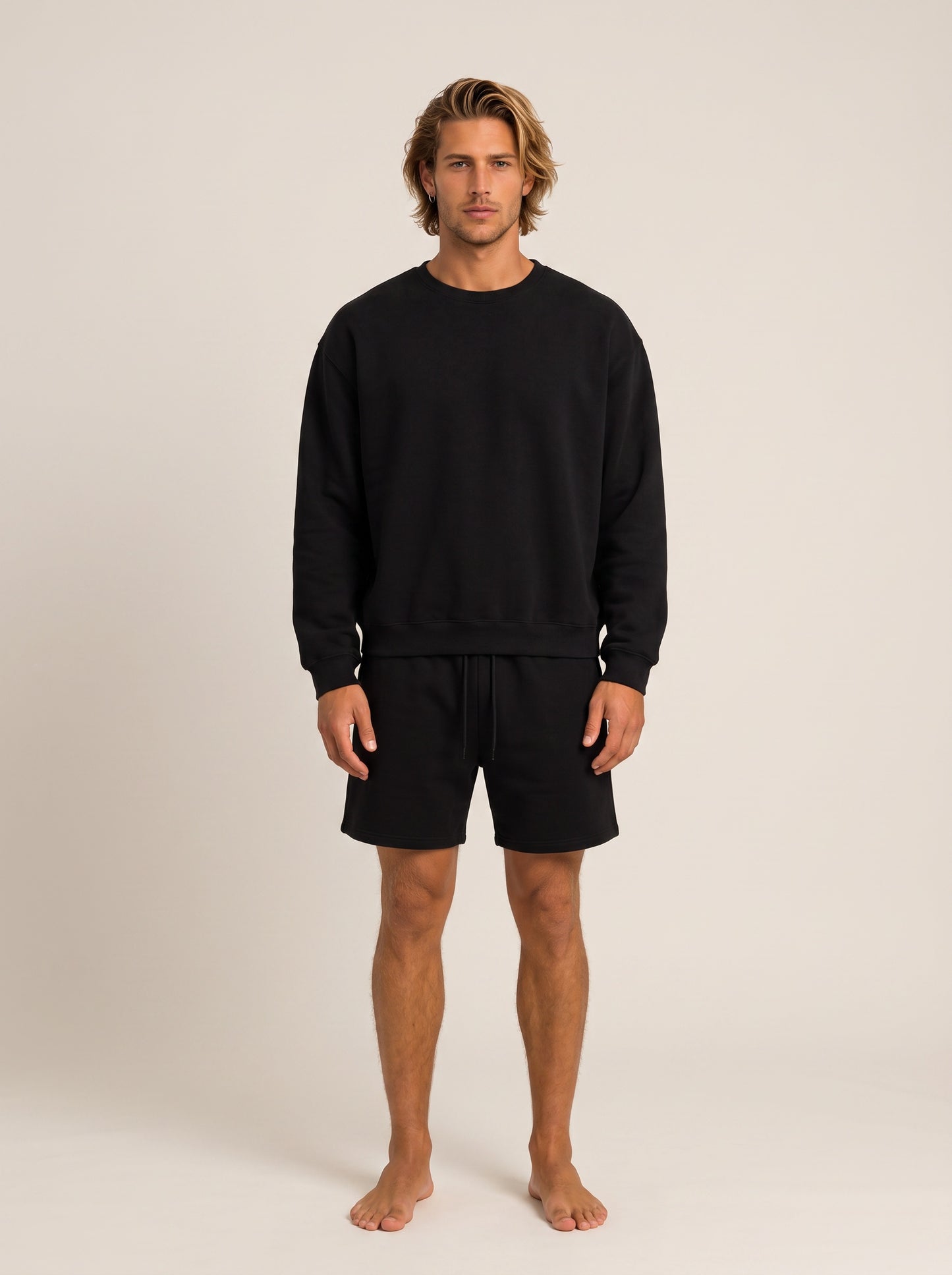 A man modeling a black crewneck sweatshirt and matching black shorts against a plain, warm off-white studio background.