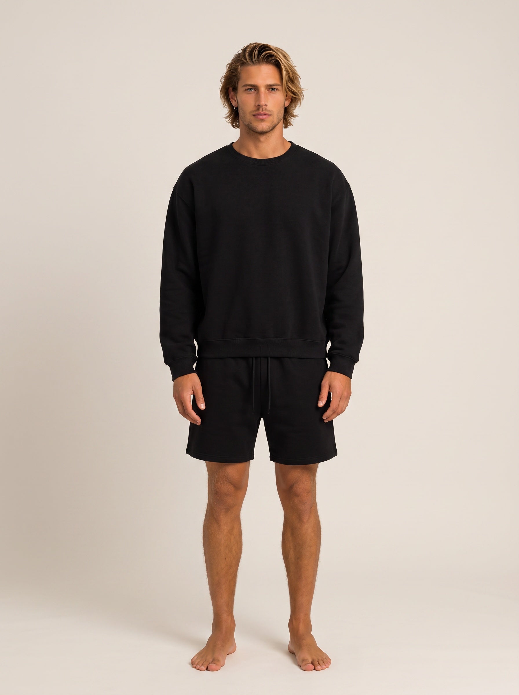 A man modeling a black crewneck sweatshirt and matching black shorts against a plain, warm off-white studio background.