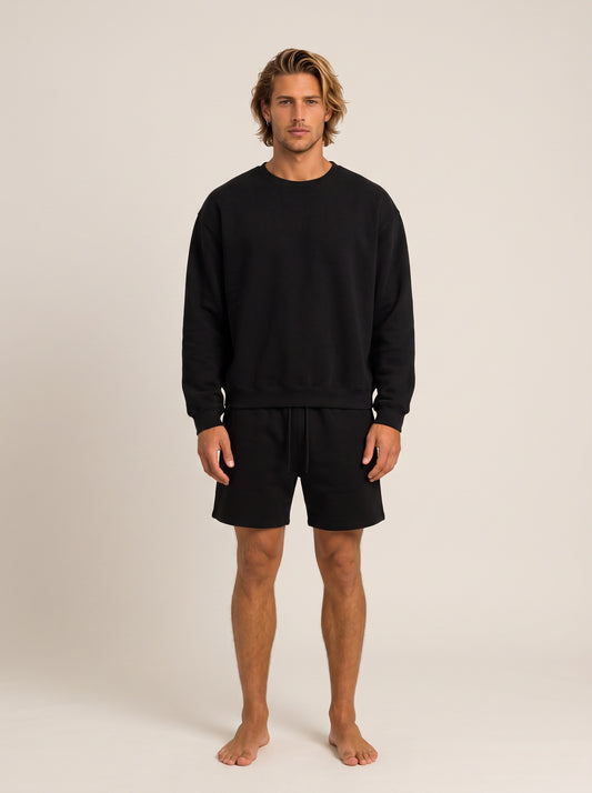 A man modeling a black crewneck sweatshirt and matching black shorts against a plain, warm off-white studio background.