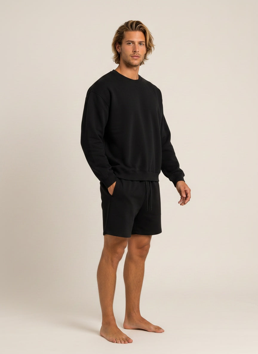 A man modeling a black crewneck sweatshirt and matching black shorts against a plain beige studio background.