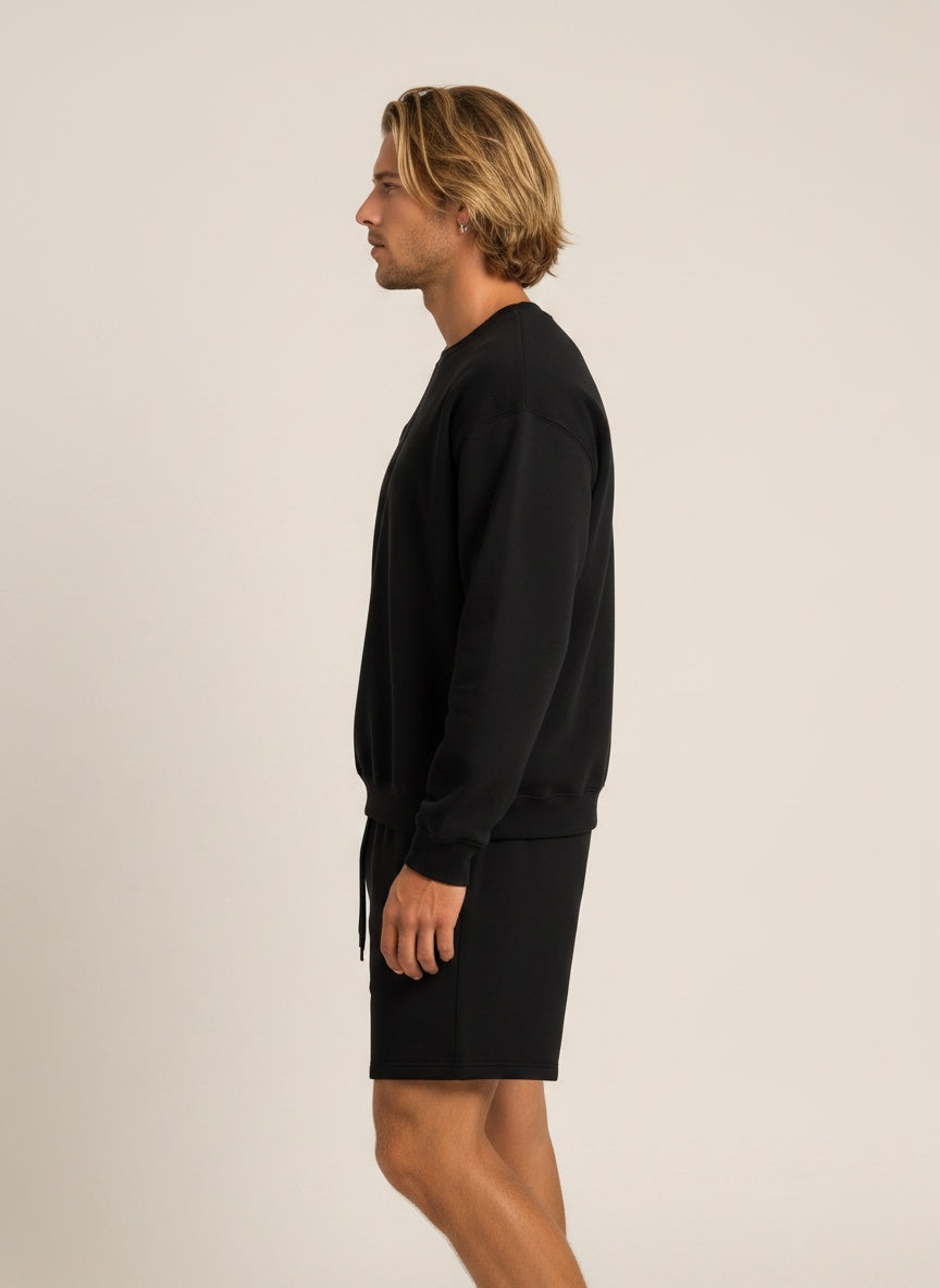 A man in profile wearing a black crewneck sweatshirt and matching black shorts against a plain light beige studio background.