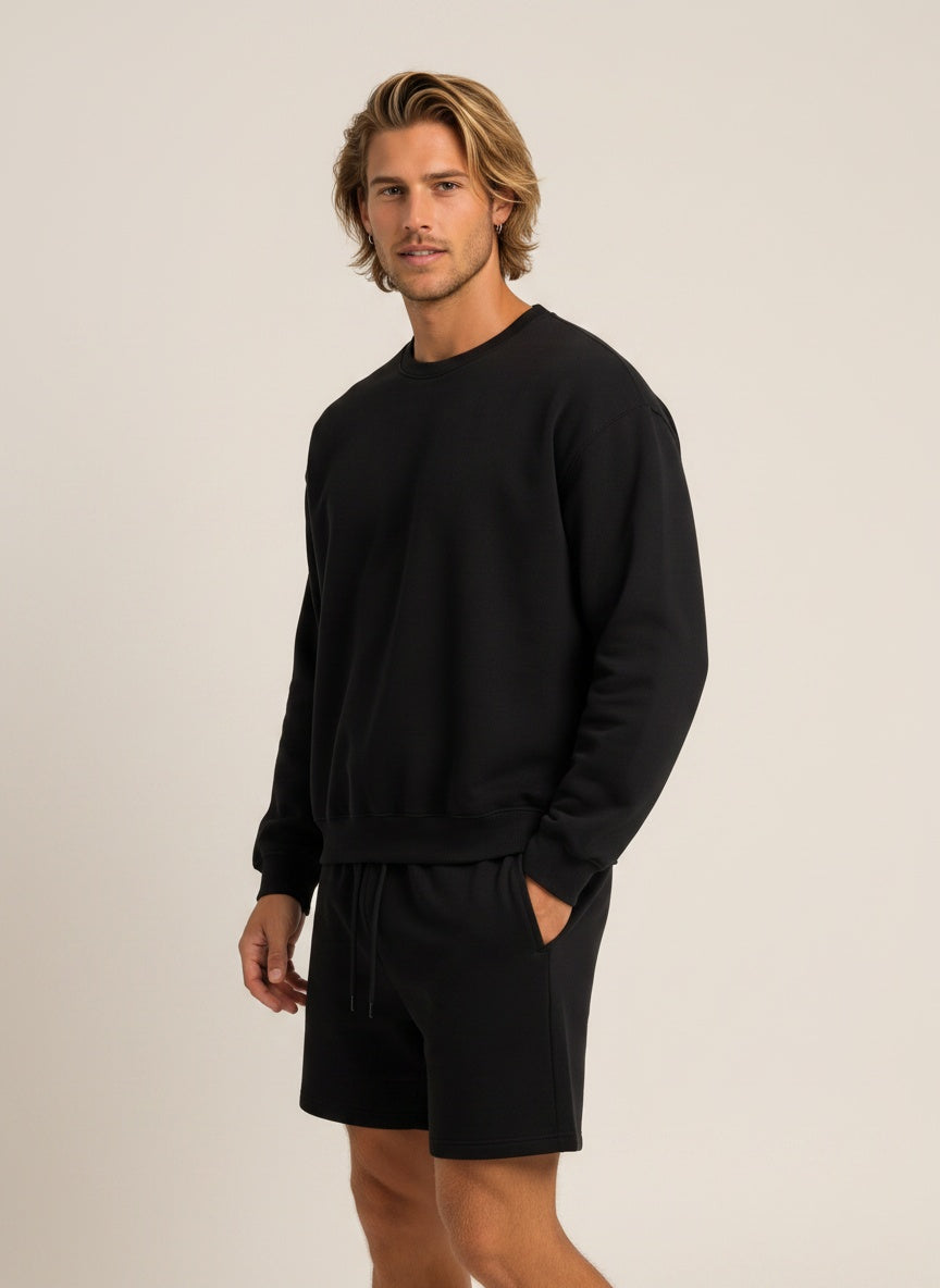 A man wearing a black crewneck sweatshirt and matching black shorts, standing against a plain light beige studio background.