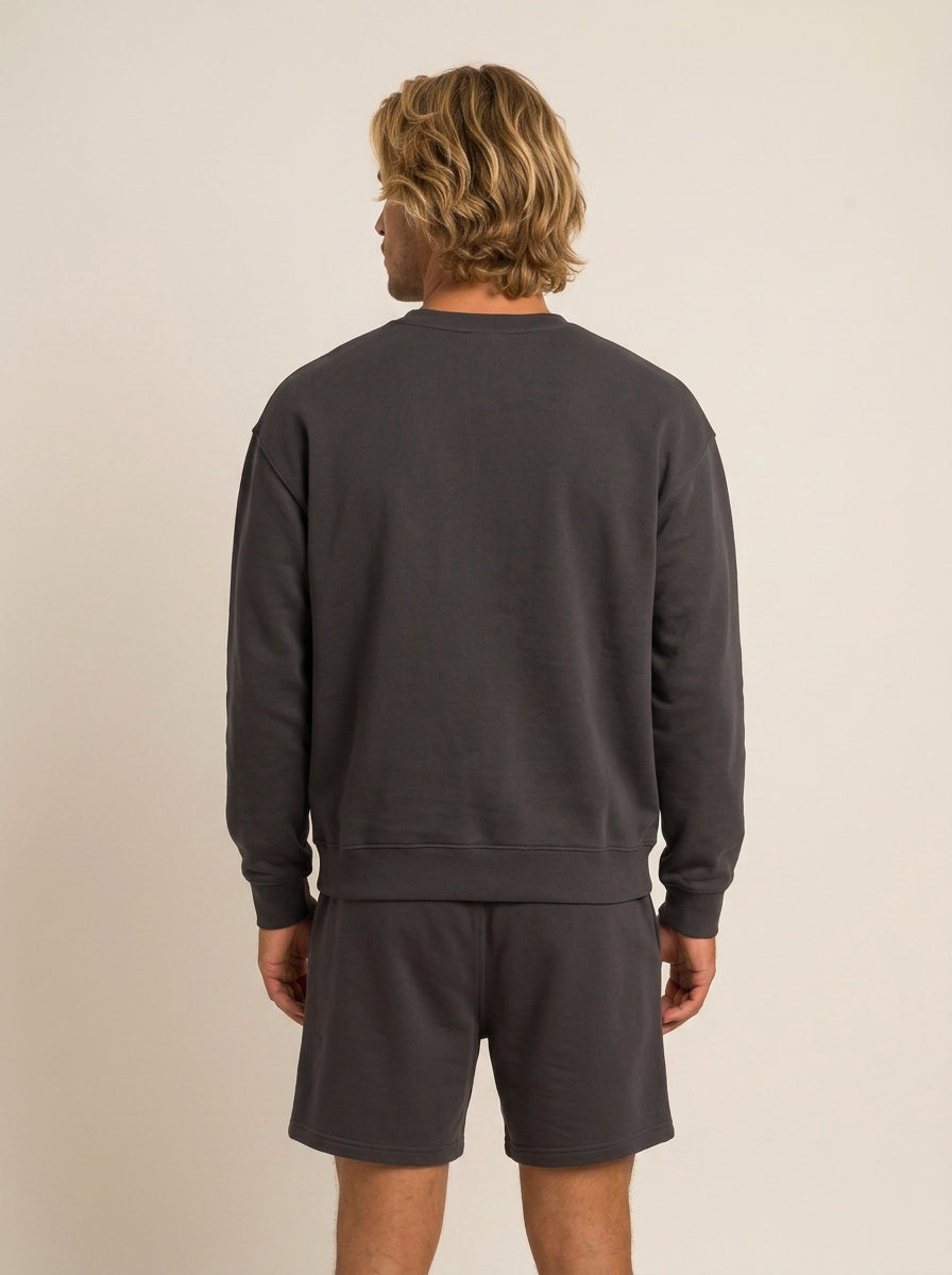 A man viewed from behind wearing a dark charcoal gray crewneck sweatshirt and matching shorts against a plain beige background.