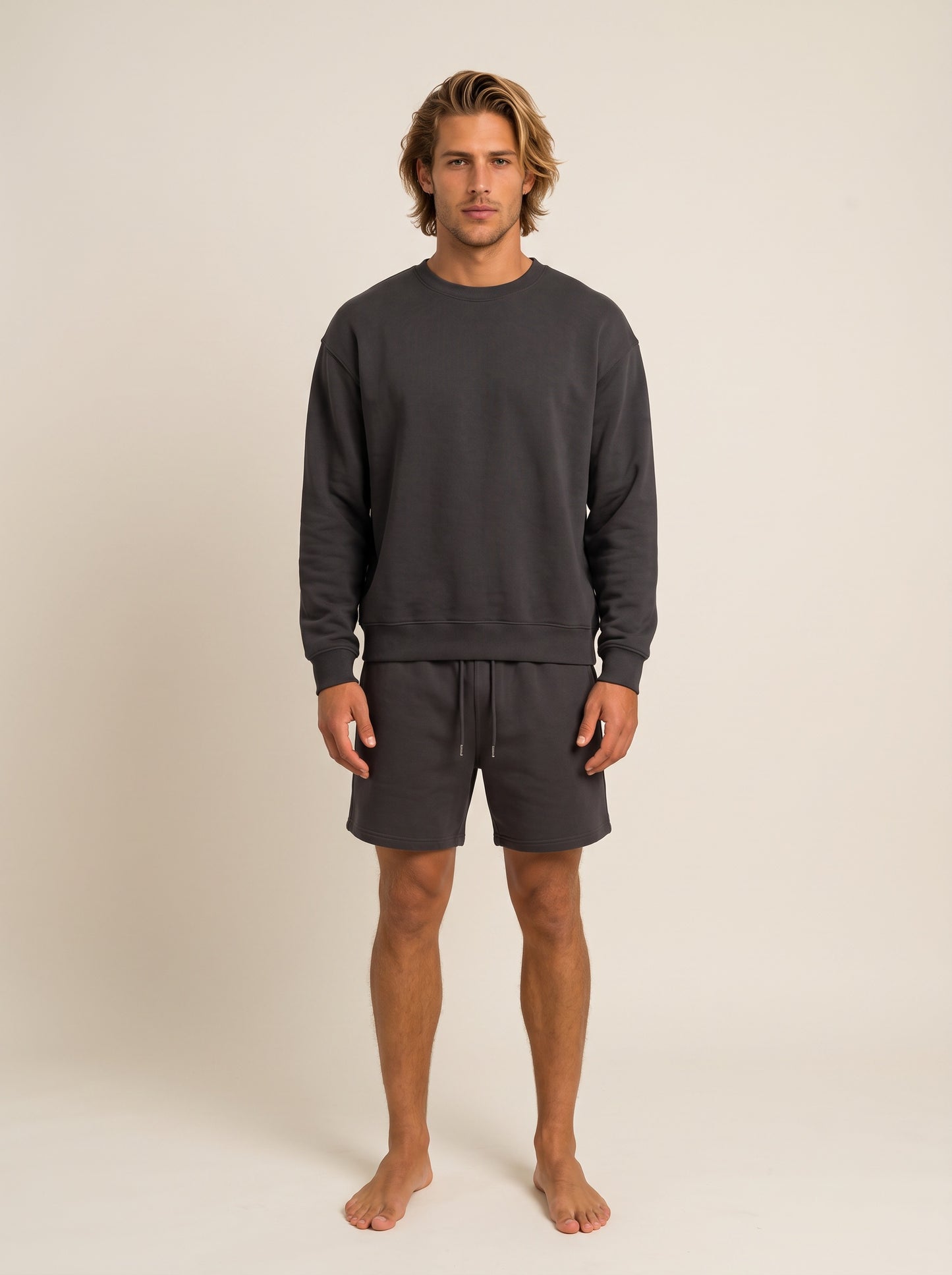 A man modeling a dark gray crewneck sweatshirt and matching dark gray drawstring shorts against a plain beige studio background.