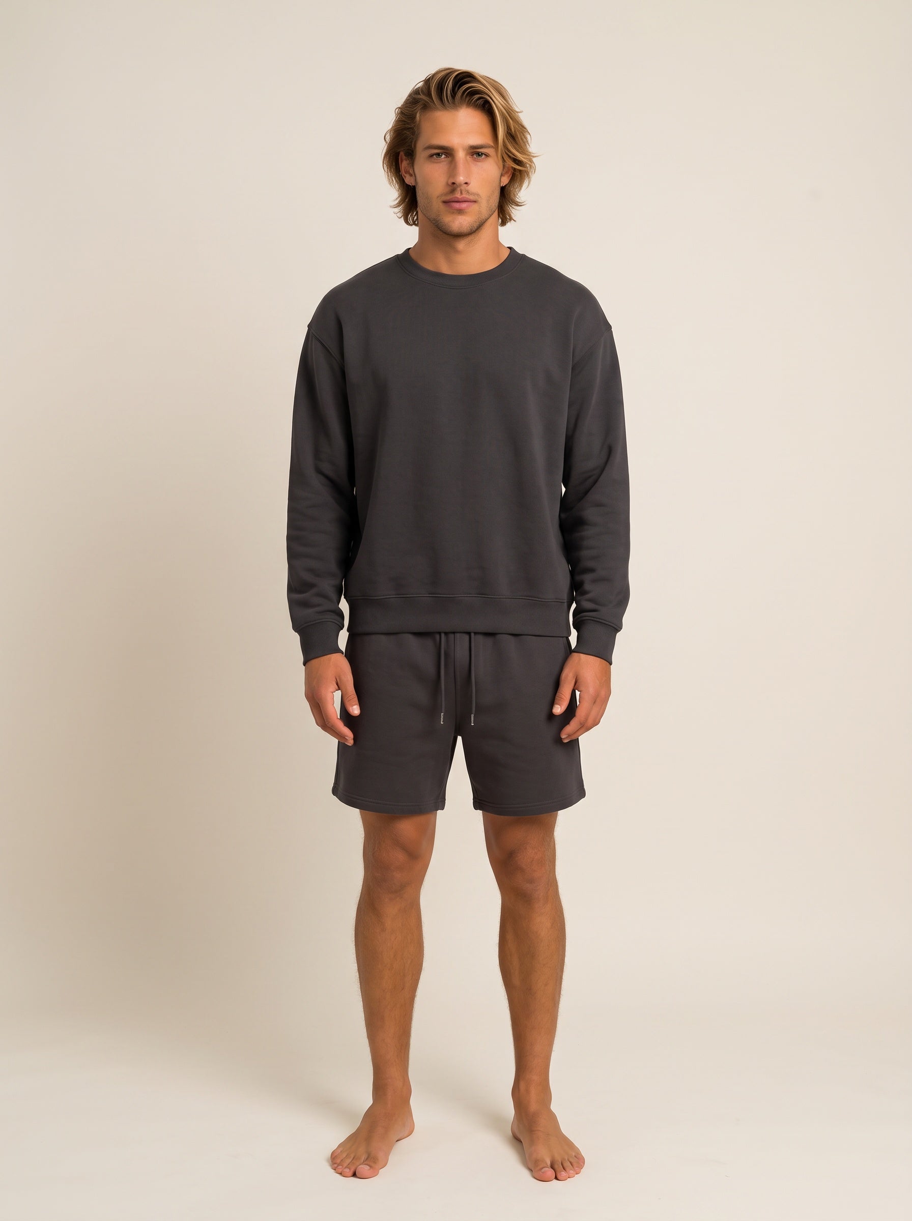 A man modeling a dark gray crewneck sweatshirt and matching dark gray drawstring shorts against a plain beige studio background.