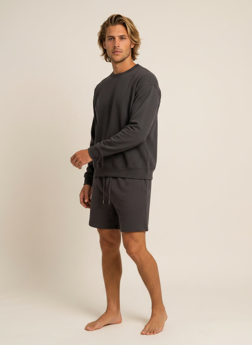 A man modeling a dark gray long-sleeve crewneck sweatshirt and matching dark gray drawstring shorts against a plain beige studio background.
