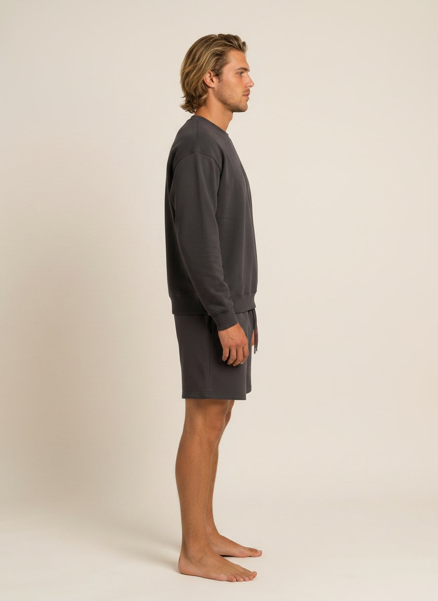 A man in side profile wearing a dark gray long-sleeve crewneck sweatshirt and matching dark gray shorts, standing barefoot against a plain beige studio background.