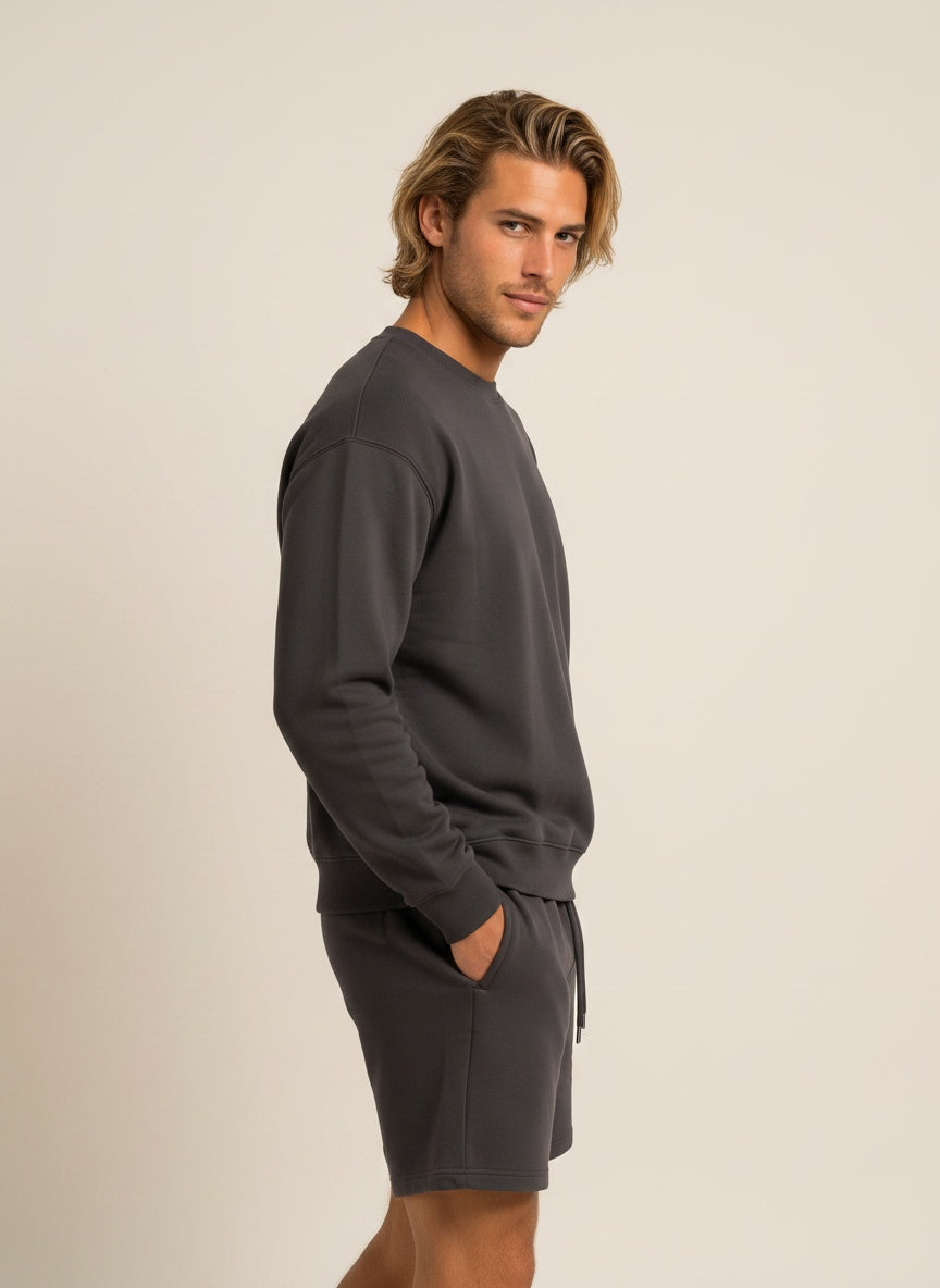 A man wearing a dark gray crewneck sweatshirt and matching shorts, shown in profile against a plain beige studio background.