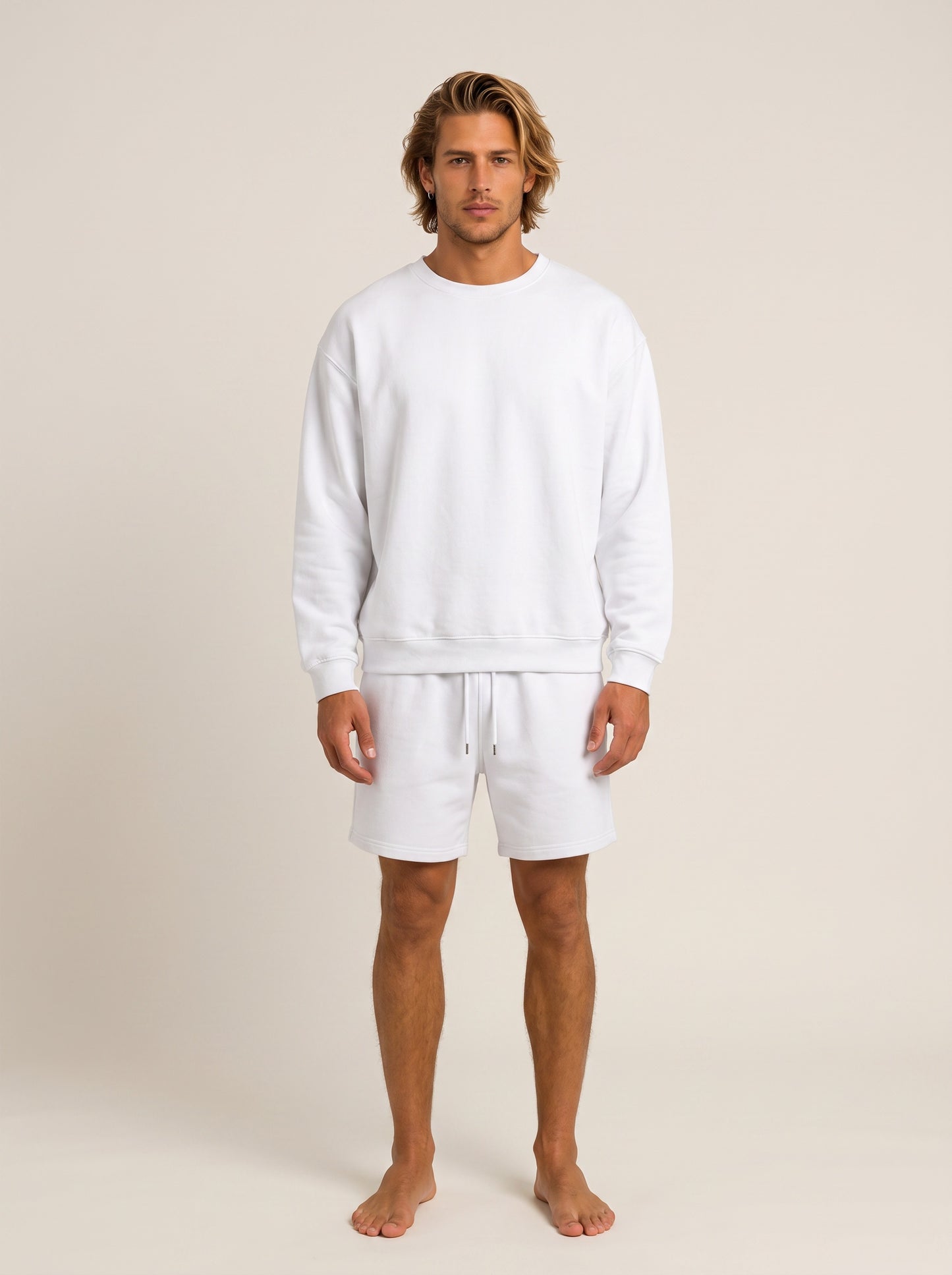 A man modeling a matching white lounge set consisting of a long-sleeve crewneck sweatshirt and drawstring shorts, standing against a solid light beige background.