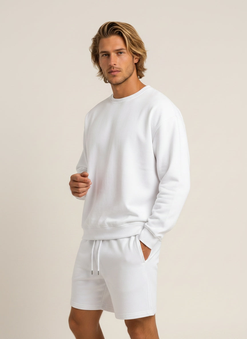 A man wearing a white long-sleeve crewneck sweatshirt and matching white drawstring shorts, standing against a plain beige studio background.