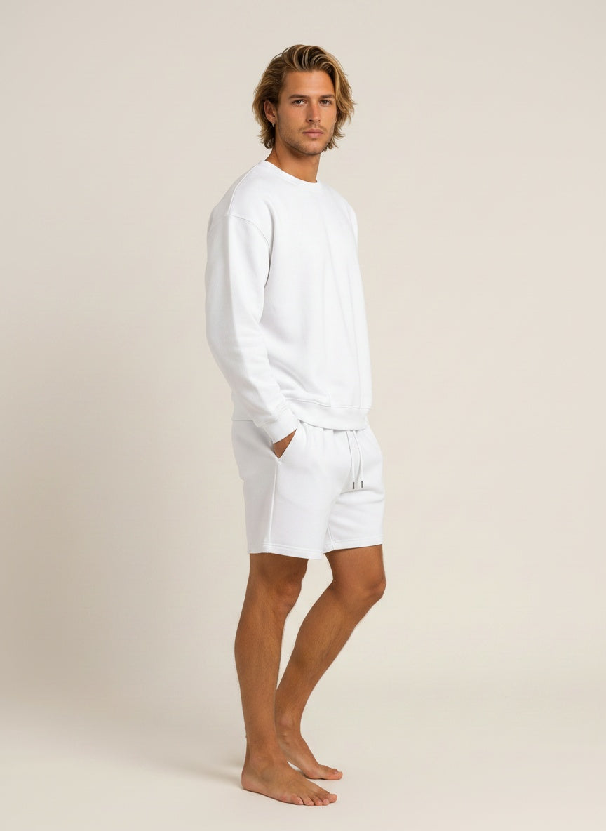 A man modeling a white long-sleeve crewneck sweatshirt and matching white shorts against a plain beige studio background.