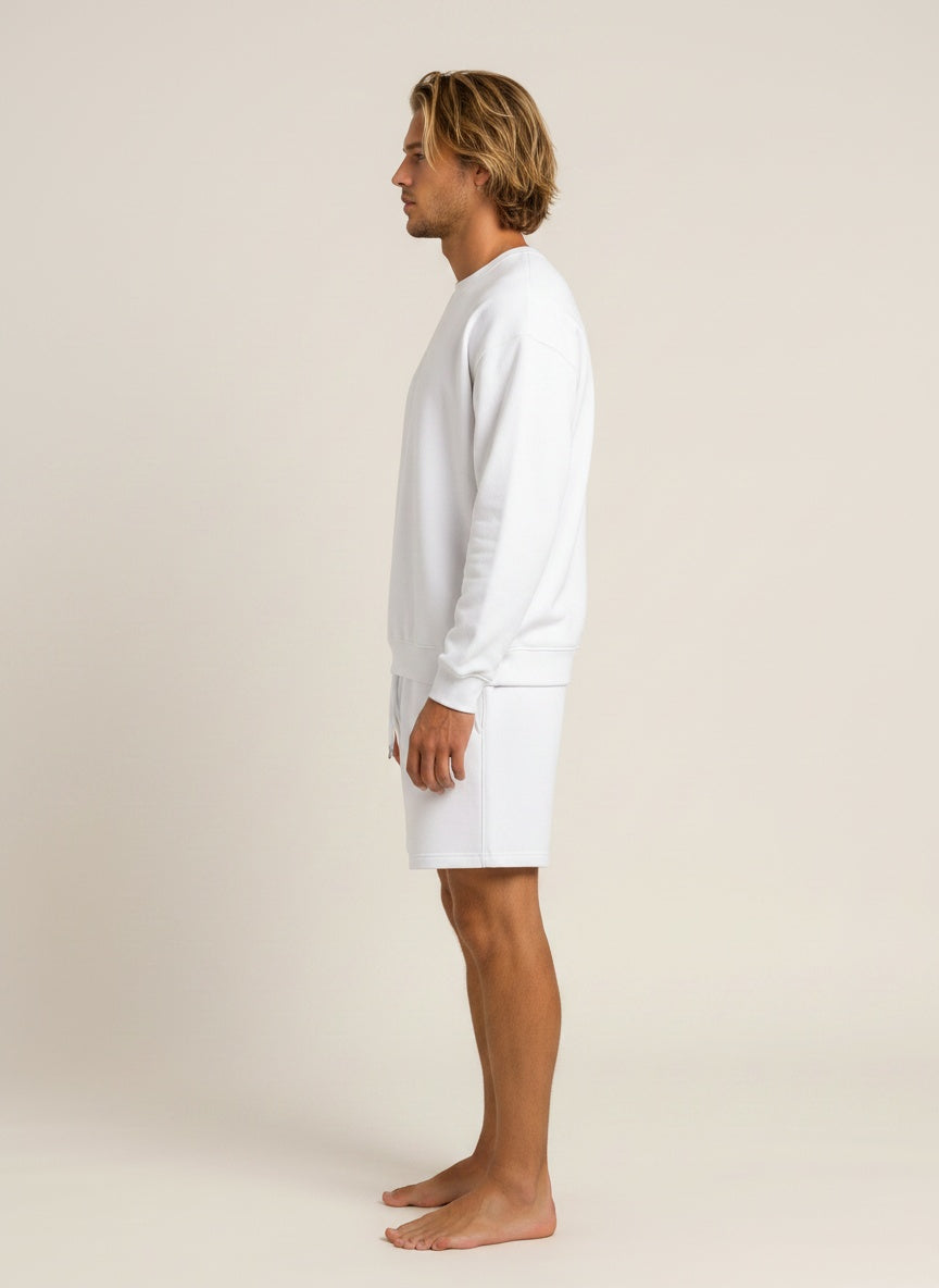 A man in profile wearing a white long-sleeve crewneck sweatshirt and matching white shorts, standing barefoot against a plain light beige studio background.