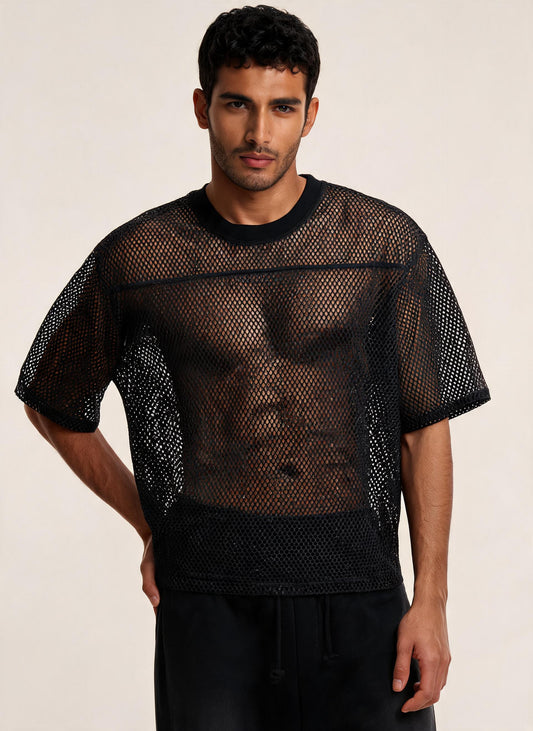 A man wearing a black short-sleeve mesh shirt over a black undershirt, paired with black pants, against a plain light beige studio background.