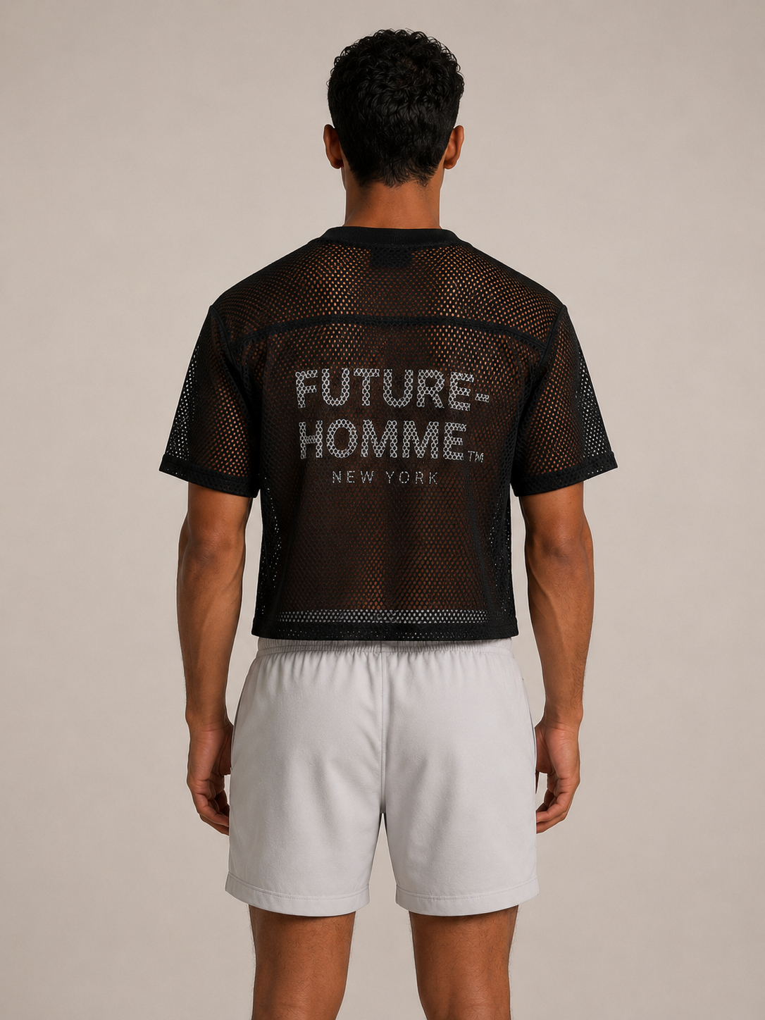 A man viewed from the back wearing a black mesh short-sleeve crop top with 'FUTURE-HOMME NEW YORK' printed in white on the back, paired with light gray shorts, against a plain beige studio background.