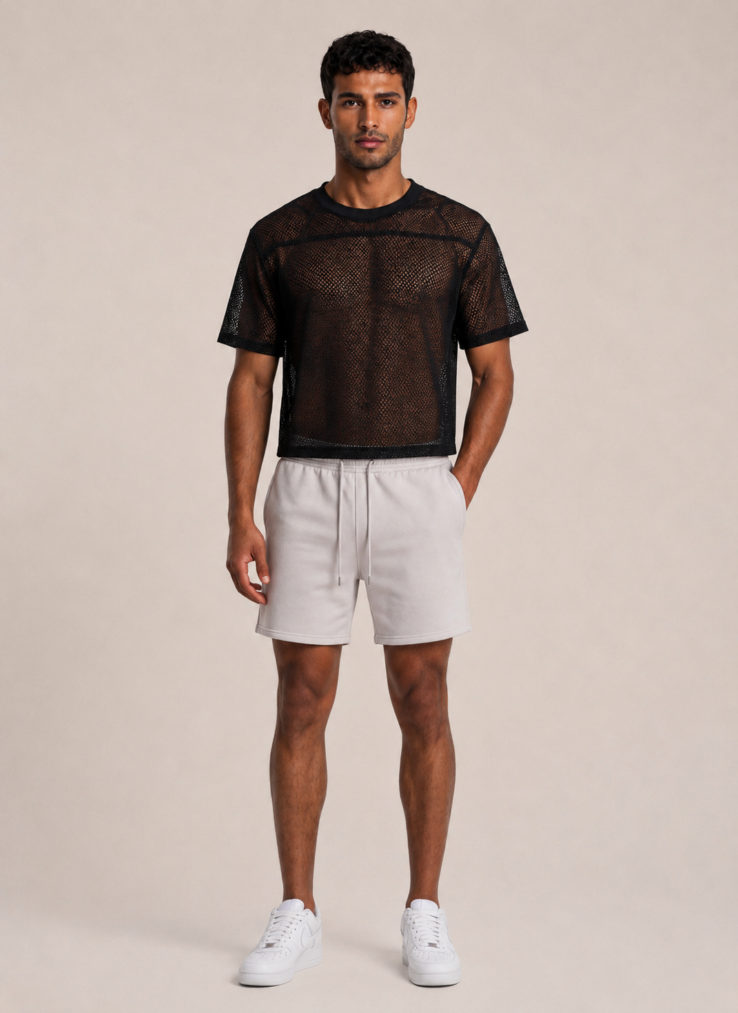 A man modeling a black sheer mesh short-sleeve t-shirt and off-white drawstring shorts against a solid beige studio background.