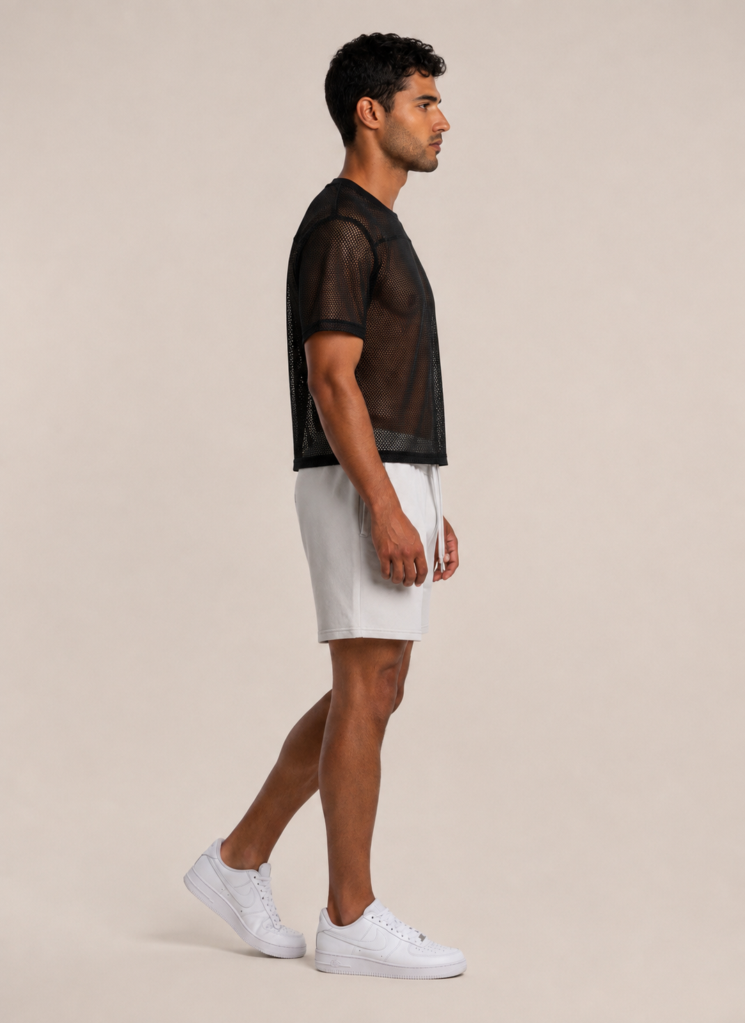 A man in side profile wearing a sheer black short-sleeve crop top, white shorts, and white sneakers, standing against a solid beige studio background.
