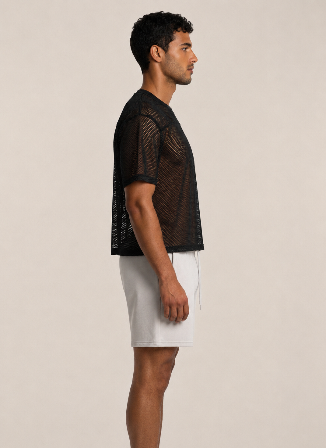 A man in profile wearing a black short-sleeve mesh cropped top and white shorts, standing against a plain light beige studio background.