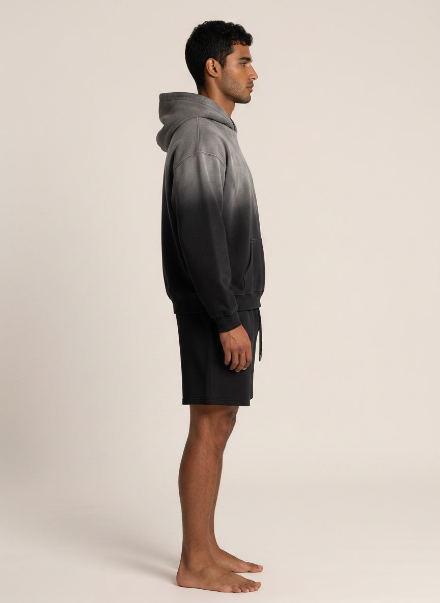 A man in profile wearing a dark gray to black gradient hoodie and black shorts, standing barefoot against a plain light beige studio background.