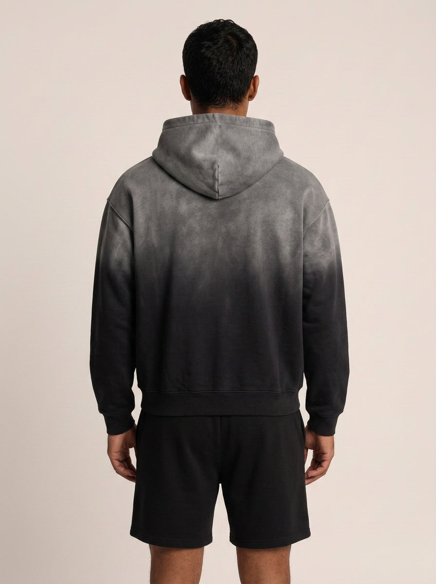 A man viewed from behind wearing a dark gray and black gradient tie-dye hoodie with black shorts, set against a plain light beige background.