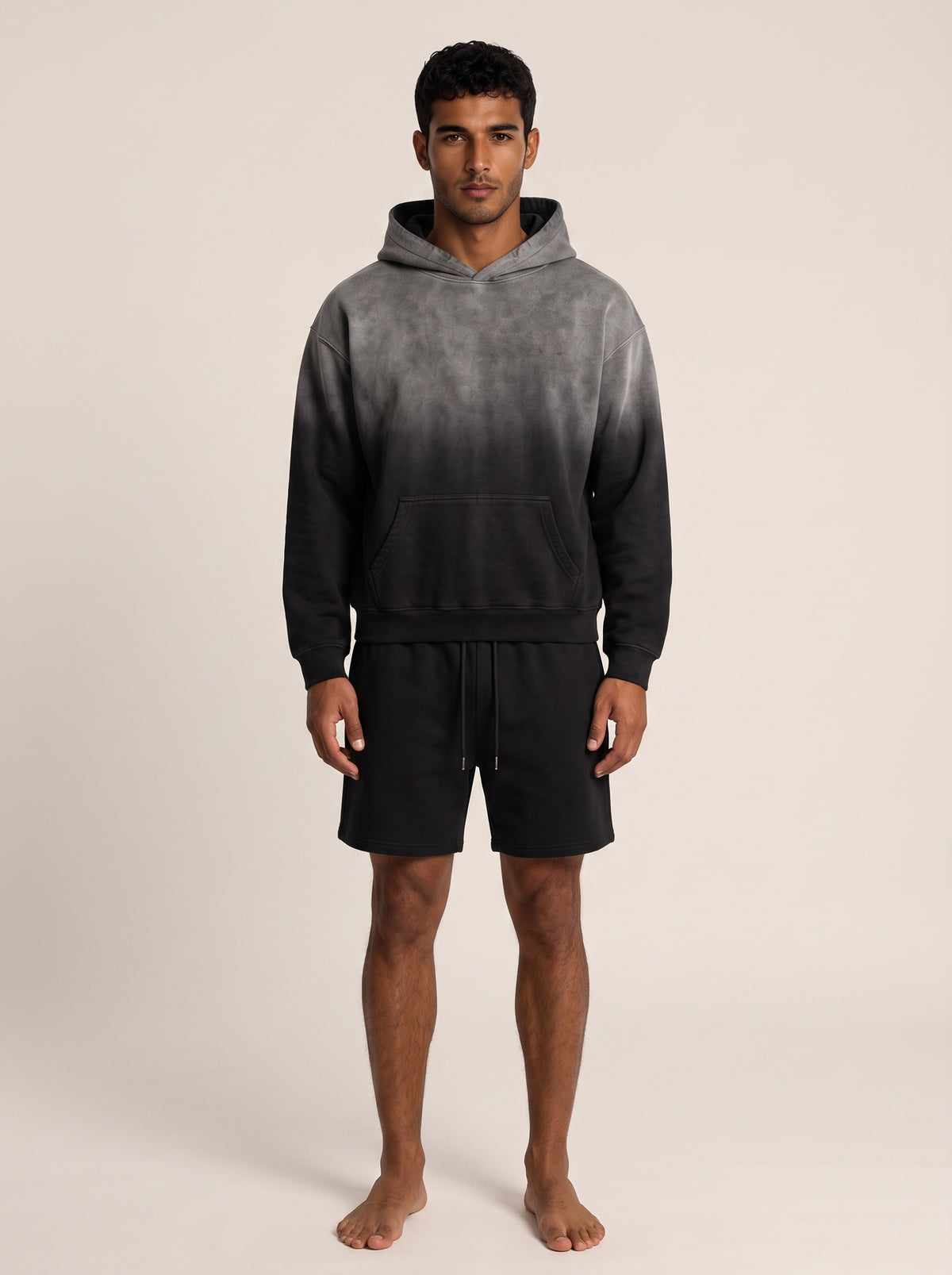 A man modeling a dark gray and black gradient hoodie with matching black drawstring shorts, standing against a solid light beige background.