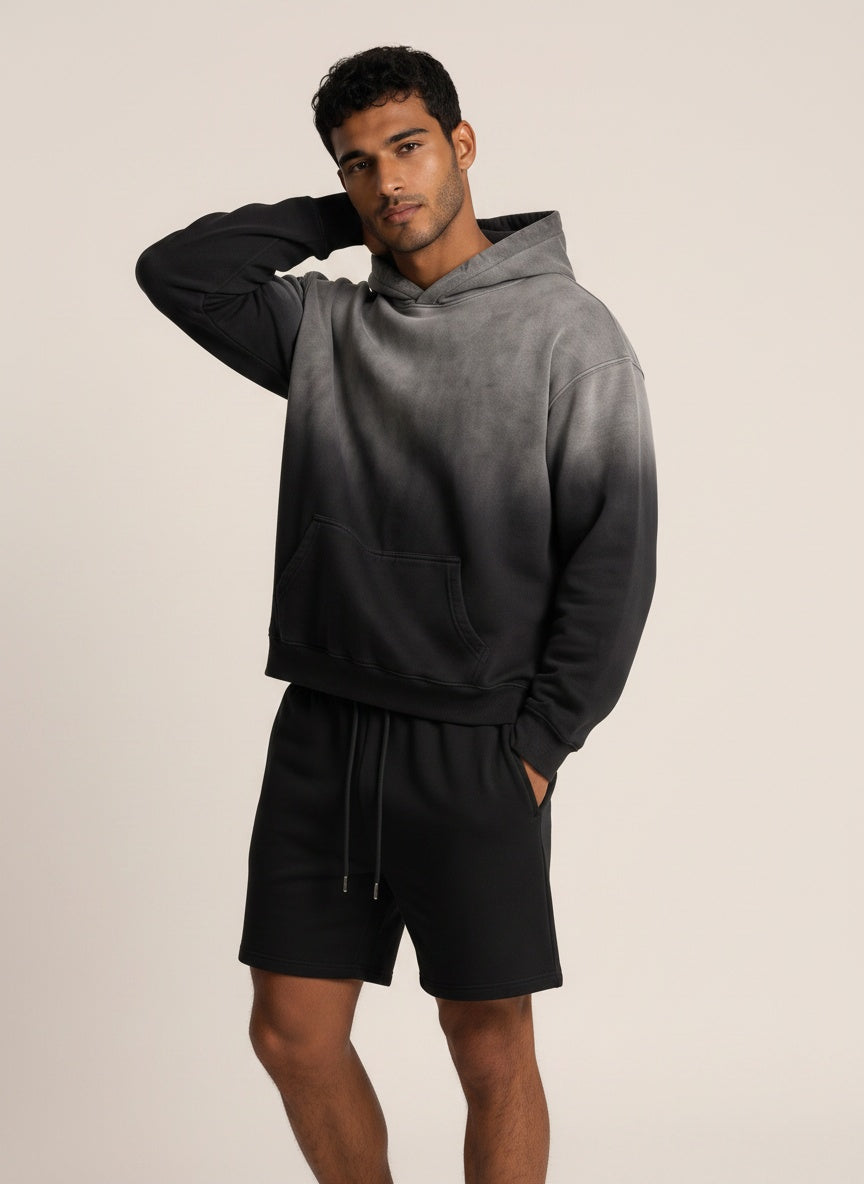 A man modeling a dark gray to black gradient hoodie with black drawstring shorts against a plain beige studio background.