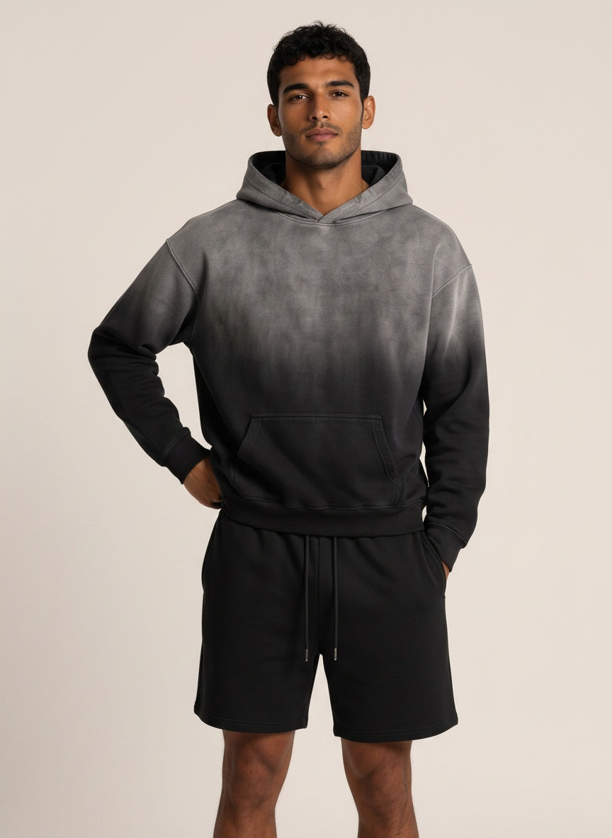 A man modeling a dark gray and black gradient hoodie with matching black drawstring shorts against a plain beige studio background.