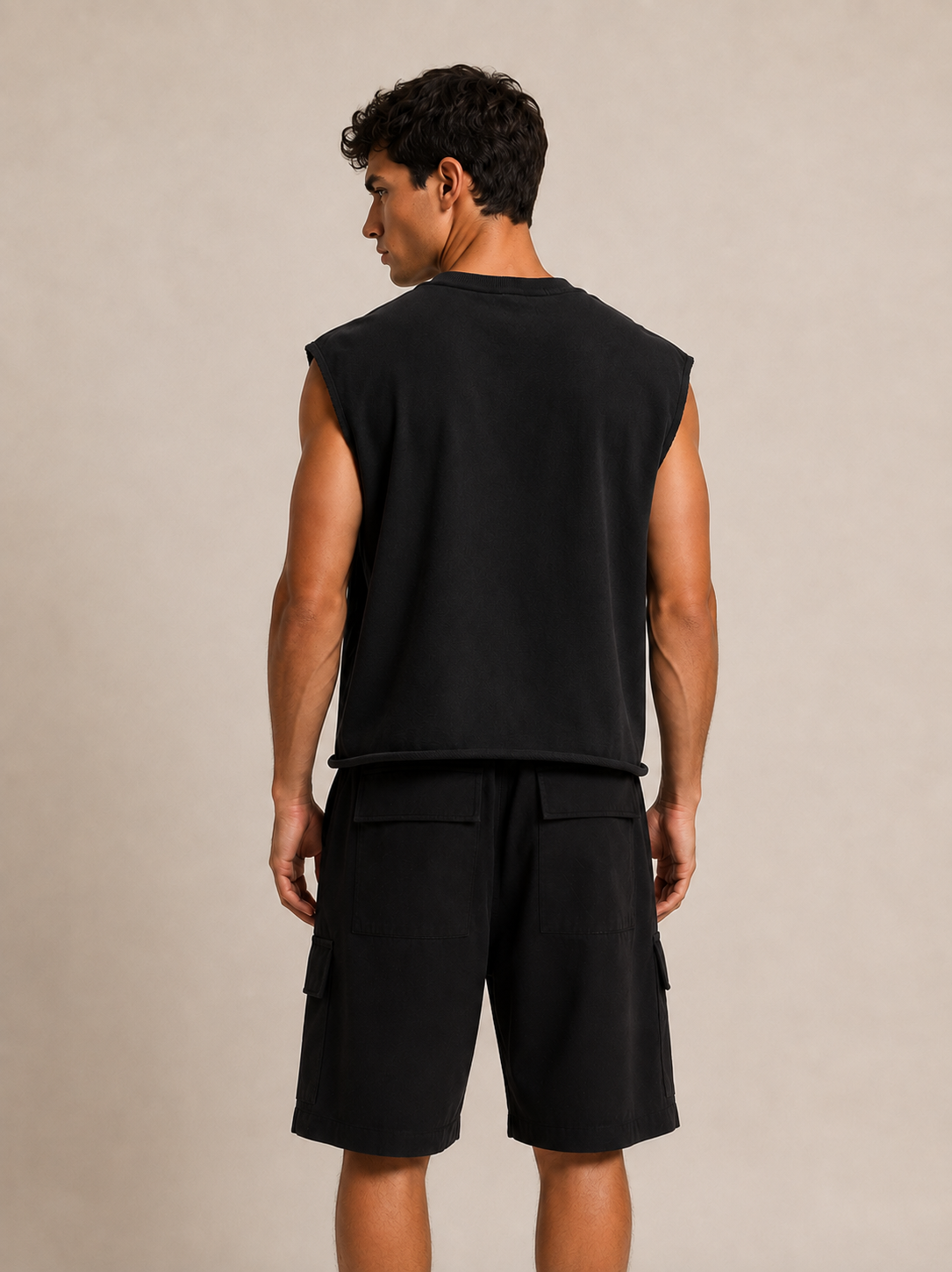 A man shown from the back wearing a black sleeveless crop top and black cargo shorts against a plain beige studio background.