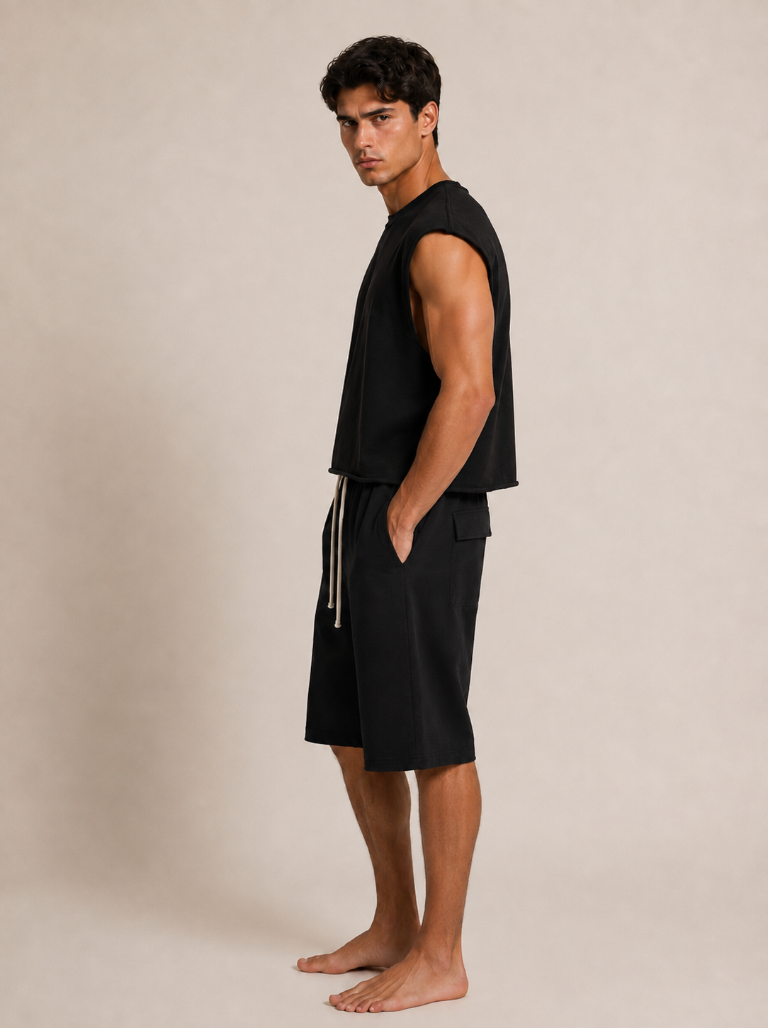 A man wearing a black sleeveless top and black shorts with a cream drawstring, standing barefoot against a plain beige studio background.