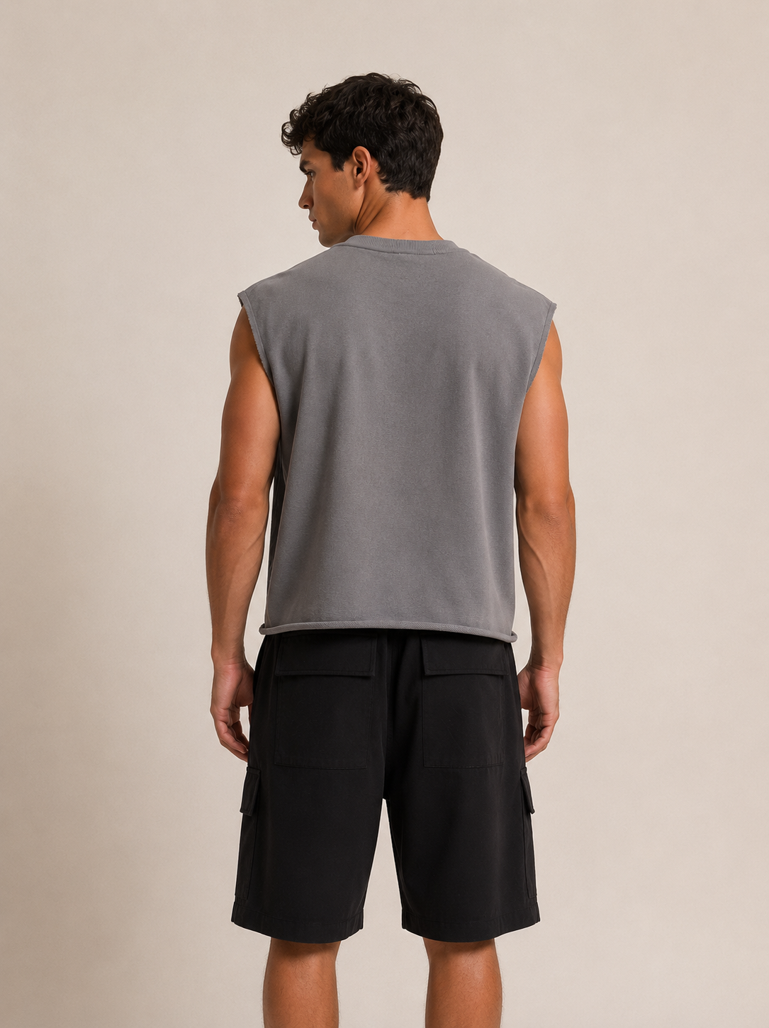 A man shown from the back wearing a gray sleeveless crop top and black cargo shorts against a plain beige studio background.