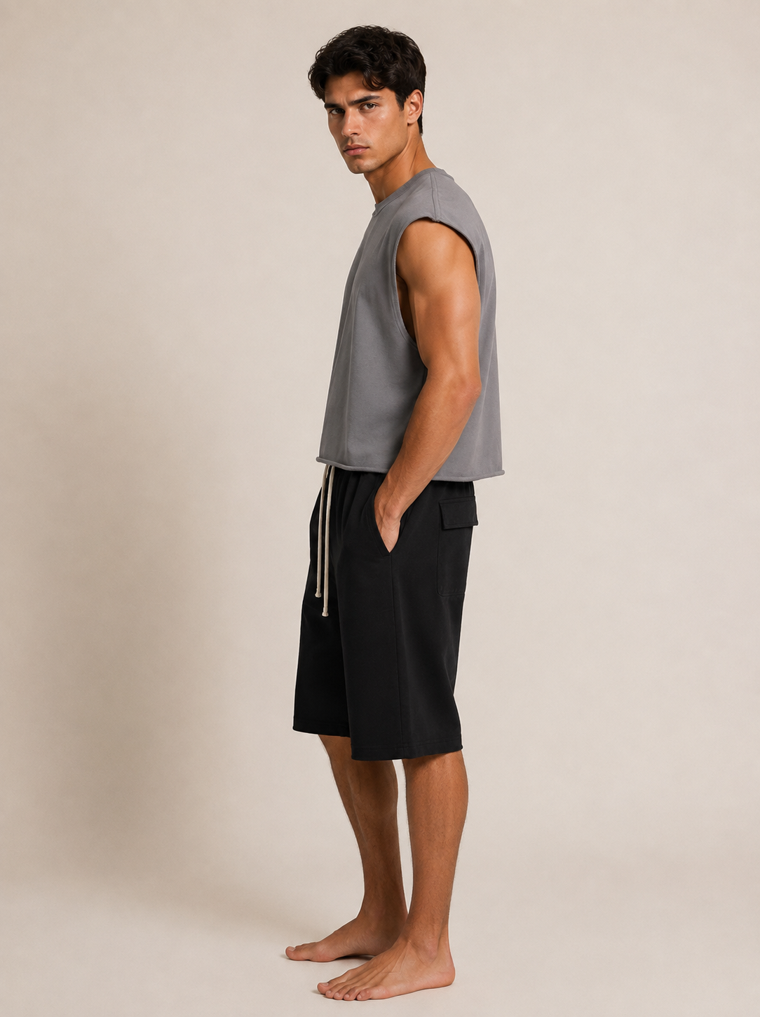 A man stands barefoot in profile wearing a sleeveless gray top and black shorts against a plain beige studio background.