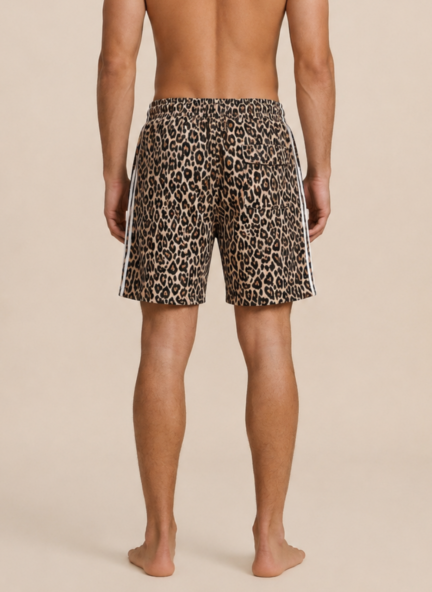 Rear view of a man wearing leopard-print swim trunks with white side stripes, standing barefoot against a plain beige studio background.