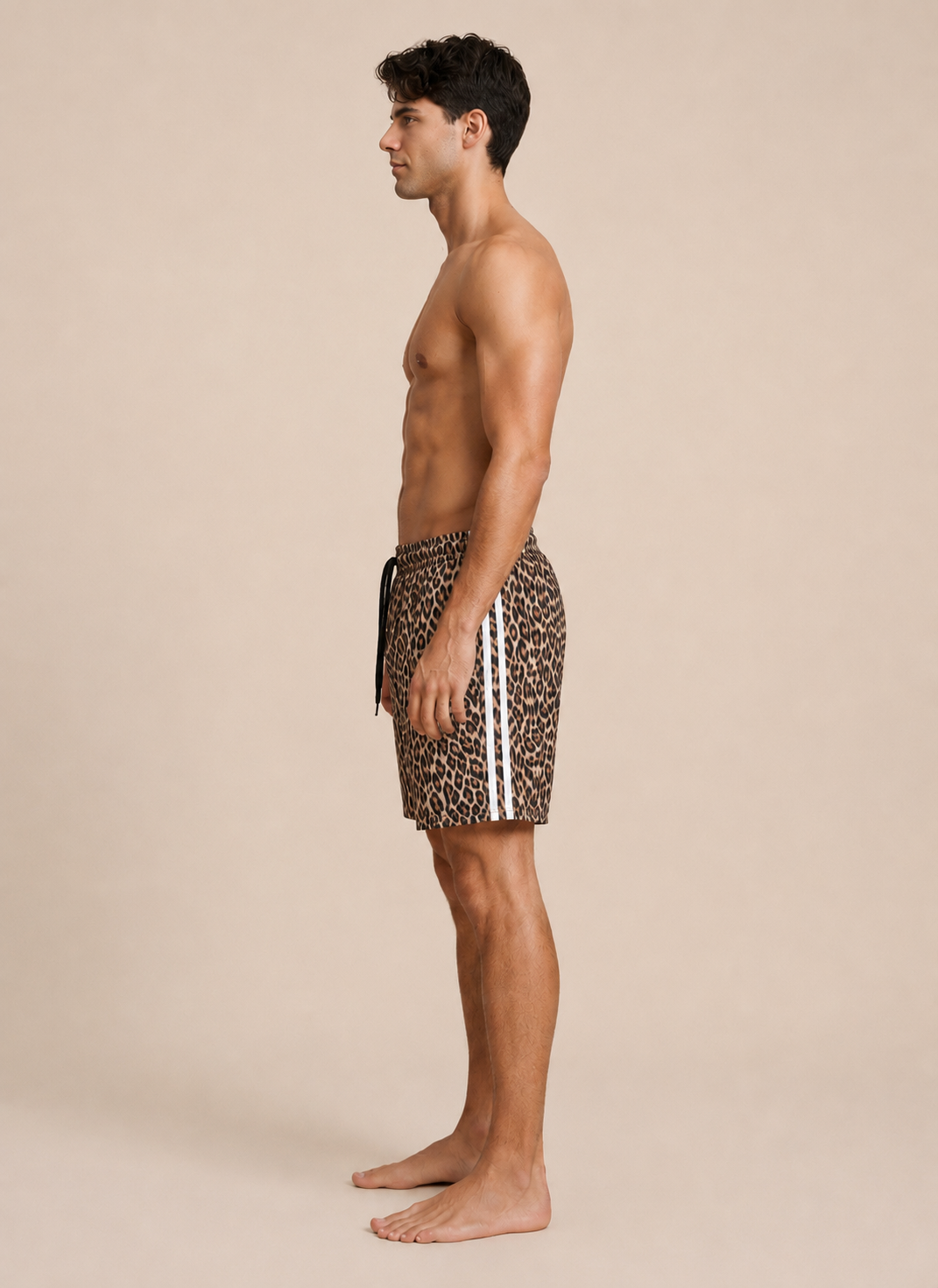 A man shown in side profile wearing leopard-print swim trunks with white side stripes, standing barefoot against a plain beige studio background.