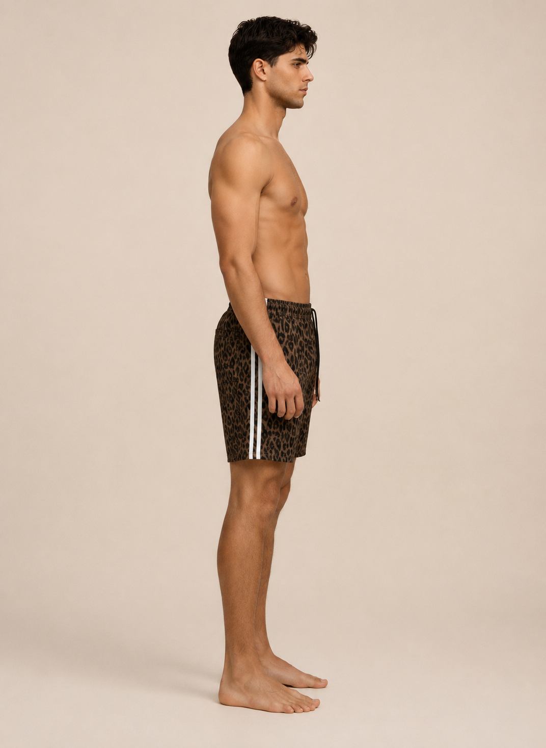 A man in side profile wearing leopard-print swim trunks with white side stripes, standing barefoot against a plain beige studio background.